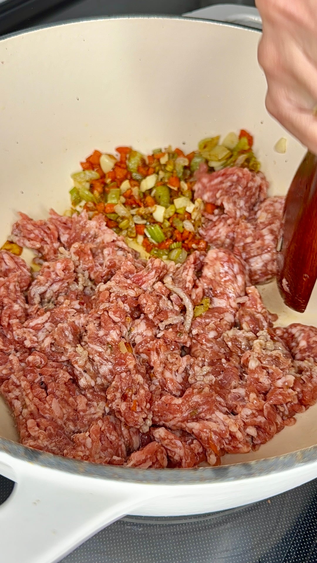 Ground meat and chopped vegetables are being sautéed in a white pot, with a hand stirring the mixture using a wooden spoon.