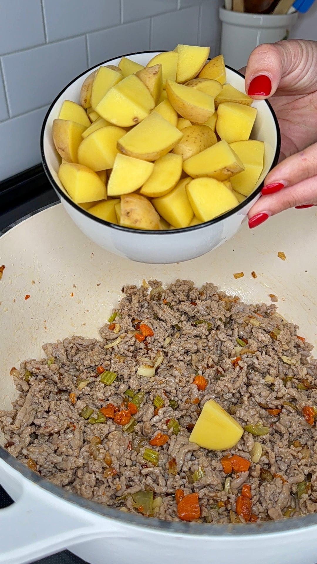 A hand holds a bowl of chopped yellow potatoes above a pot containing cooked ground meat with diced carrots and celery.