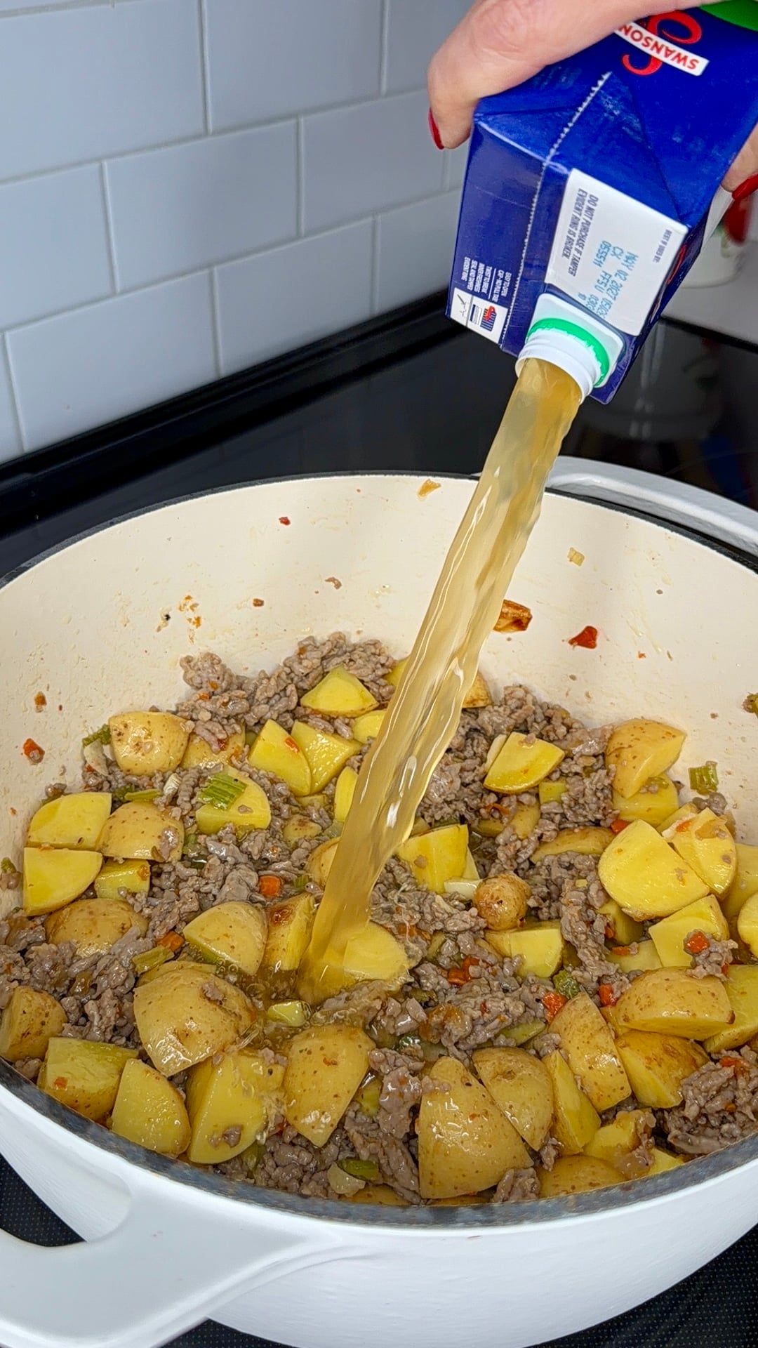 A hand pours broth from a carton into a white pot filled with browned ground meat, chopped potatoes, and vegetables on a stovetop.