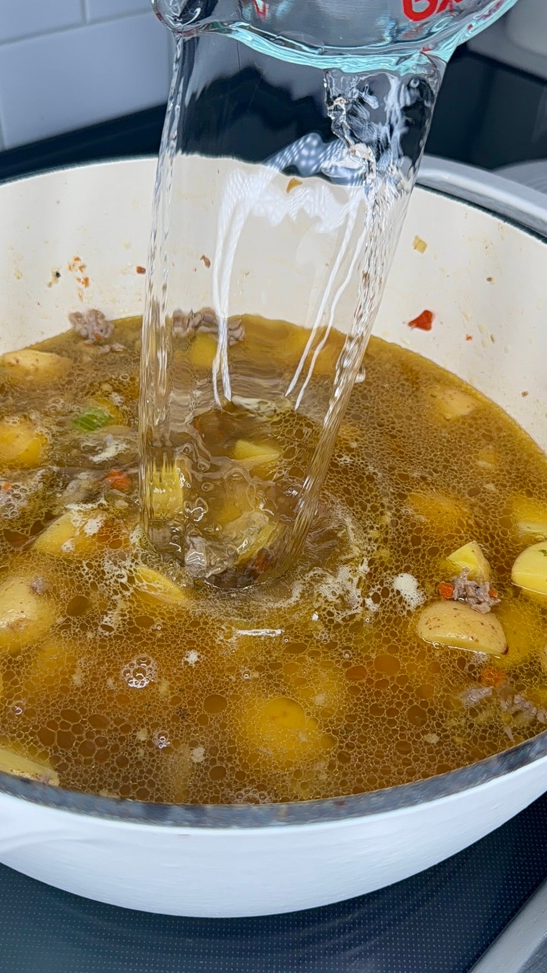 Water is being poured from a glass measuring cup into a pot of simmering stew with chunks of potatoes, vegetables, and meat.