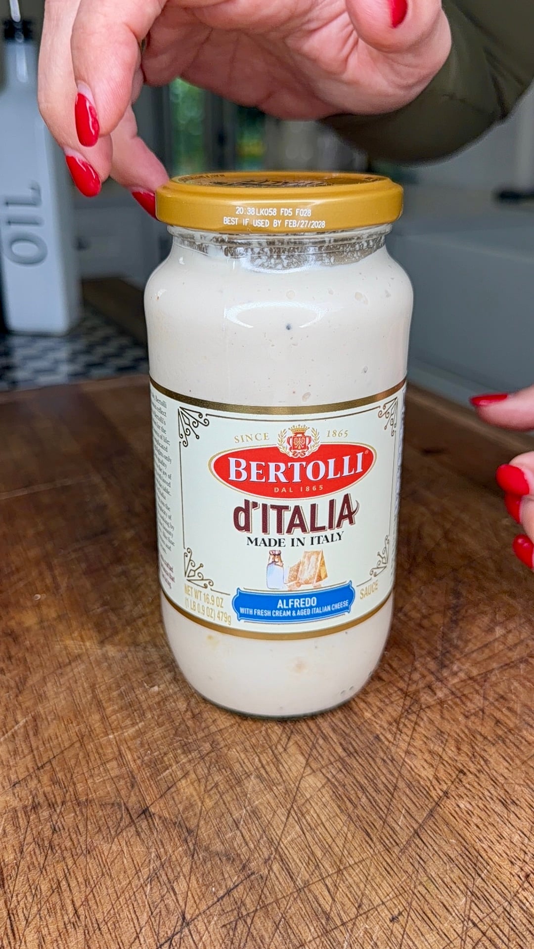 A person with red nail polish holds a jar of Bertolli dItalia Alfredo sauce on a wooden surface, ready to open it.