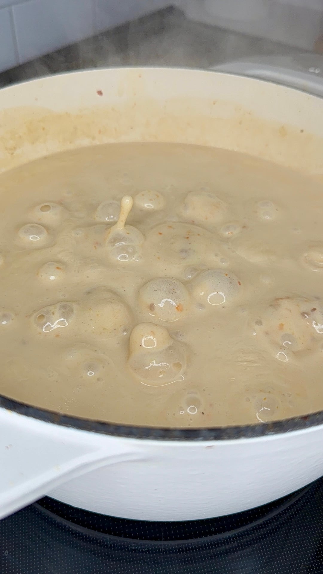 Creamy sauce with meatballs bubbling in a white pot on a stovetop, with steam rising and some splashes on the side of the pot.