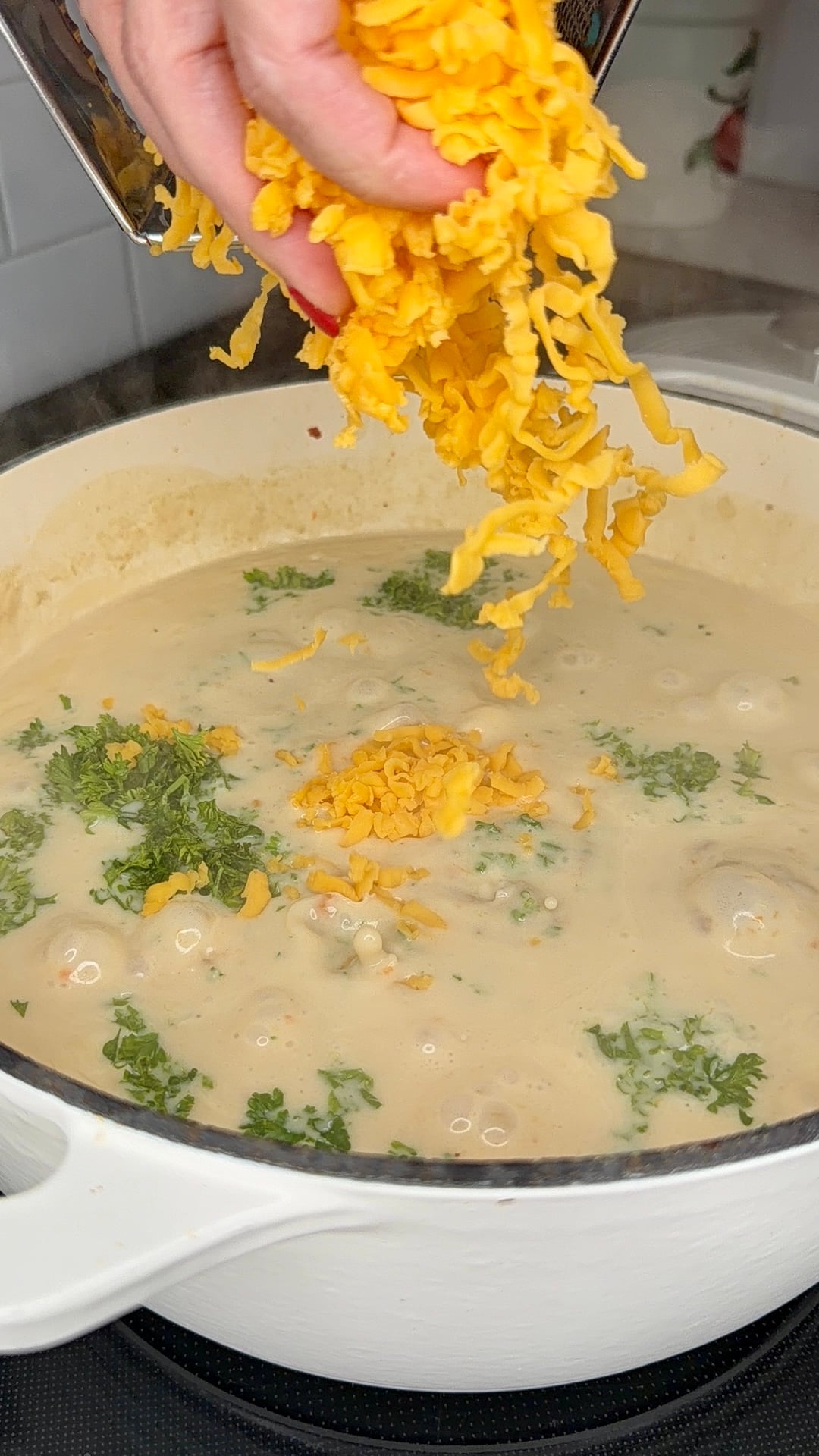A hand sprinkles shredded cheddar cheese into a pot of creamy soup with herbs on top, cooking on a stovetop in a white Dutch oven.