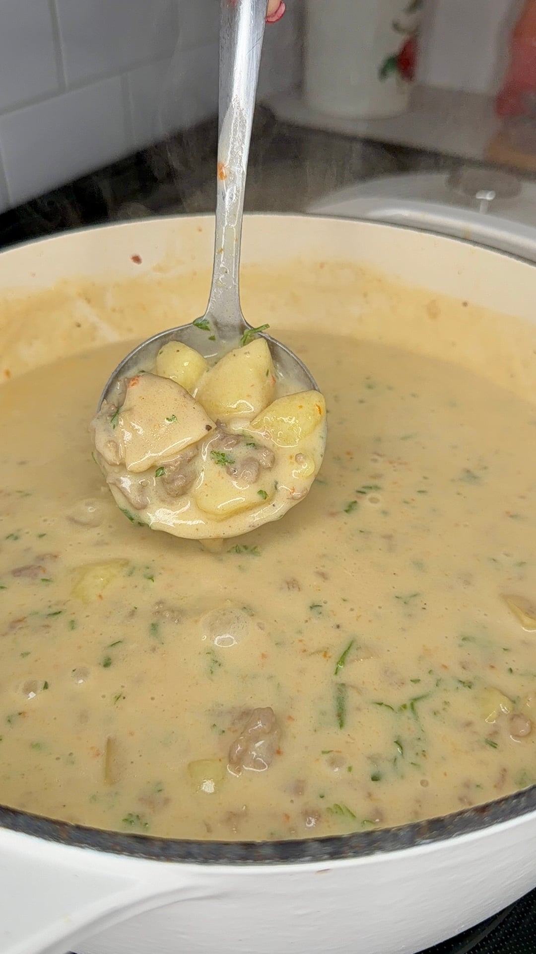 A ladle lifts creamy soup with potatoes, meat, and herbs from a white pot, showing a hearty, thick texture. Steam rises from the pot, indicating the soup is hot and freshly cooked.