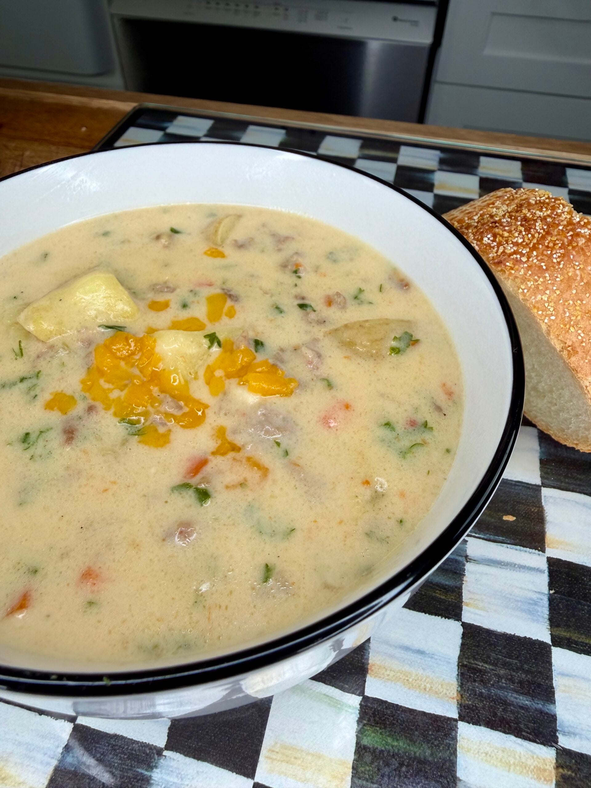 A bowl of creamy soup with herbs, vegetables, and shredded cheese on top sits next to a slice of bread on a checkered placemat.