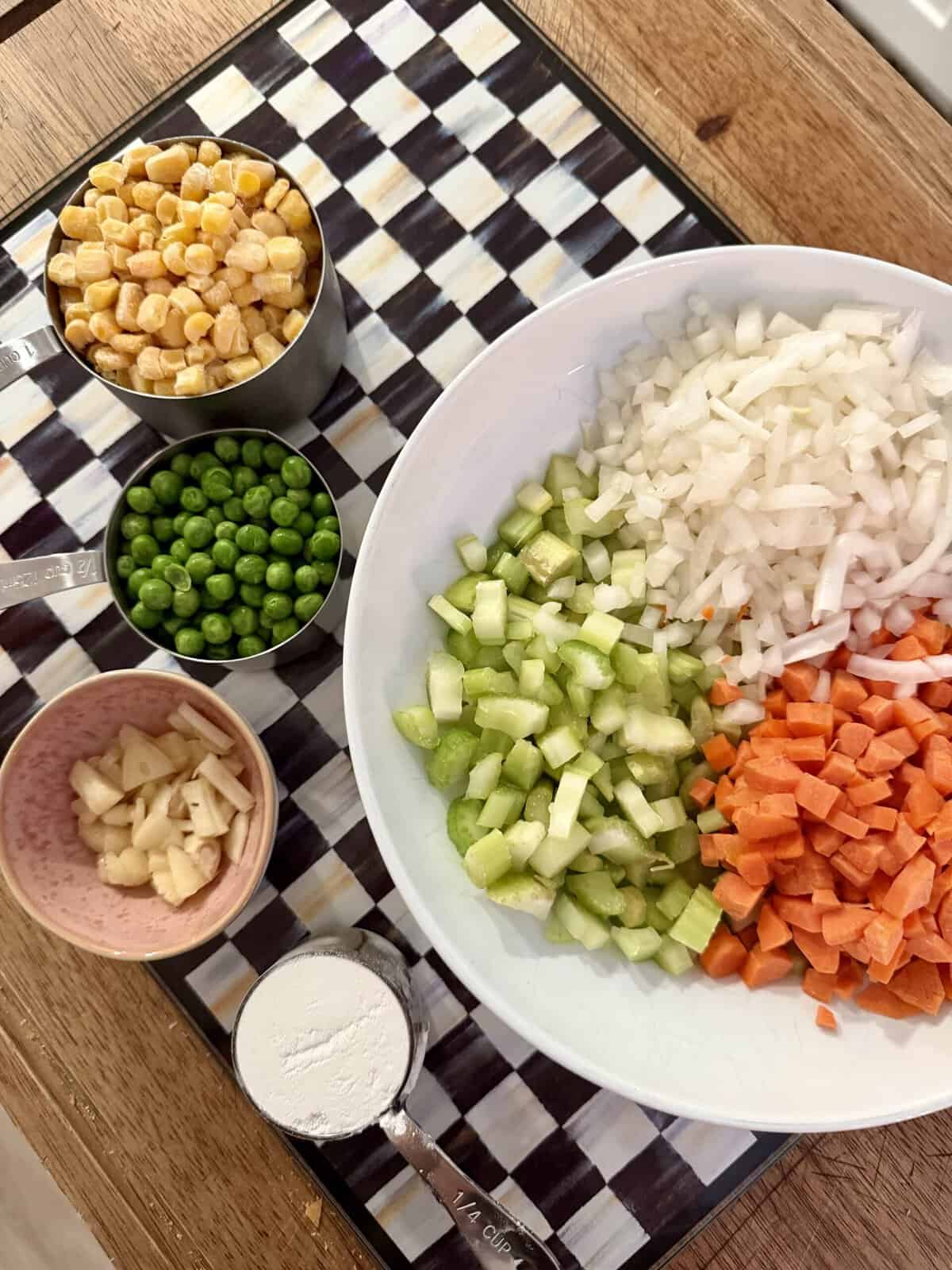 Chopped onions, celery, and carrots in a white bowl, with measured cups of corn, peas, garlic, and flour on a checkered cutting board on a wooden table.