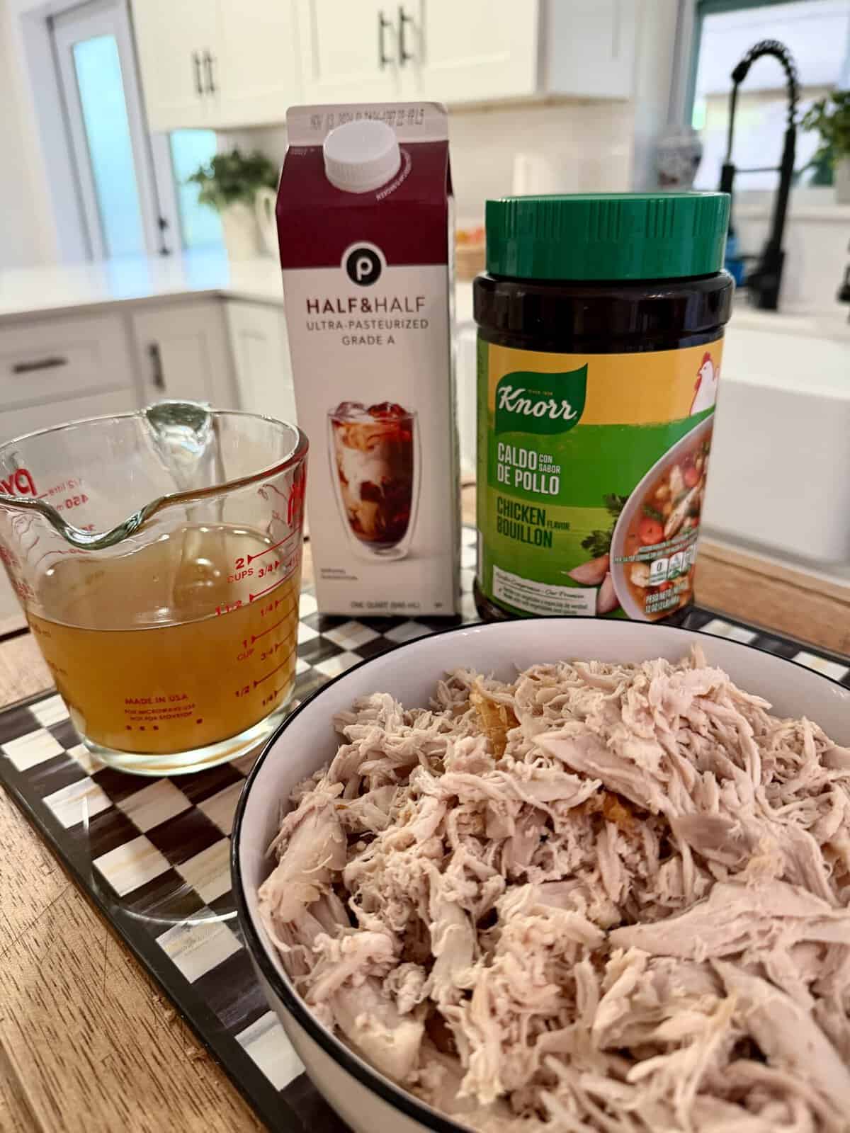 A bowl of shredded chicken sits beside a measuring cup of broth, a carton of half & half, and a jar of Knorr chicken bouillon on a kitchen counter.
