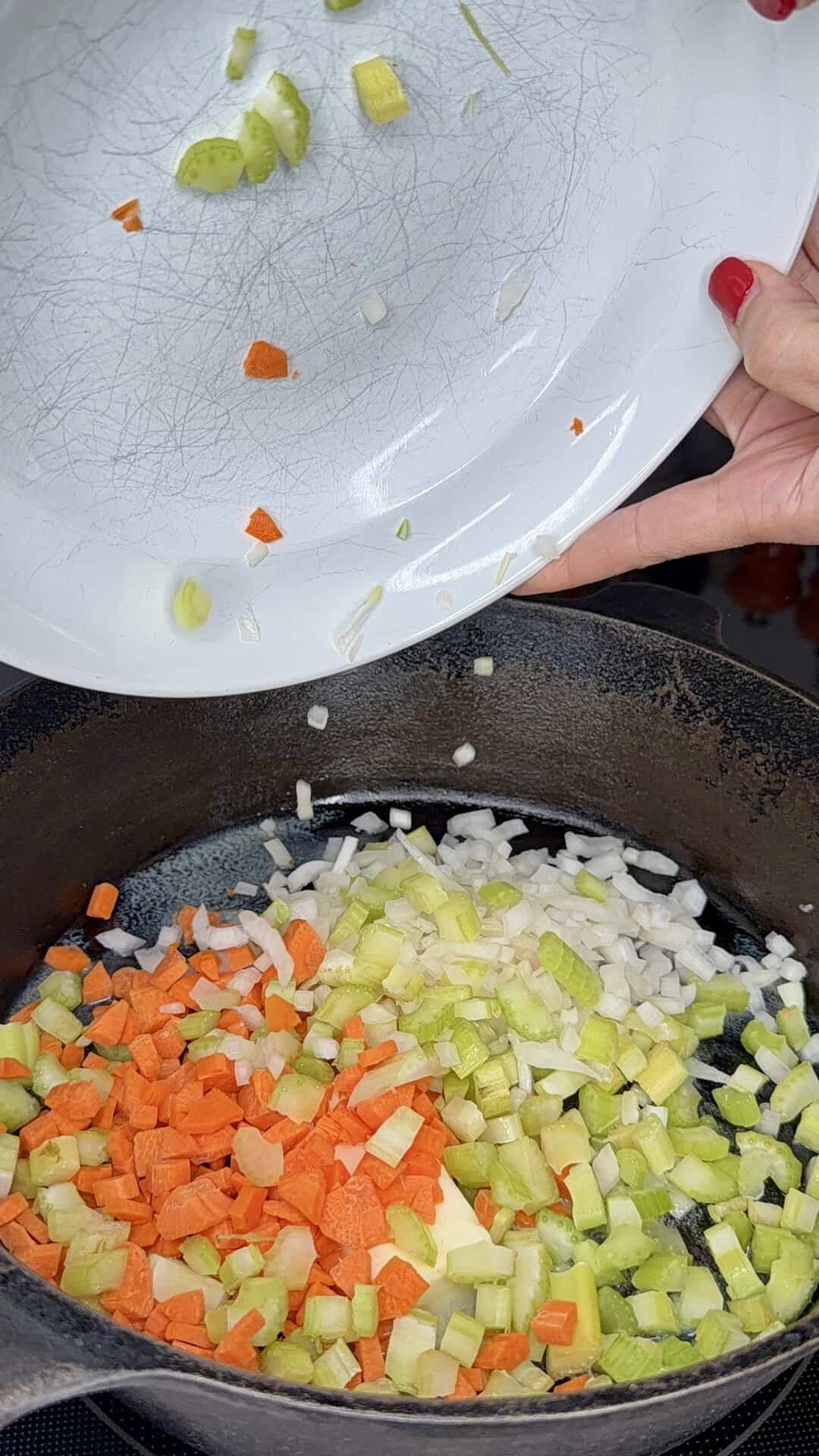 A hand pours chopped carrots, celery, and onions from a white plate into a black cast iron skillet, starting to sauté the vegetables.
