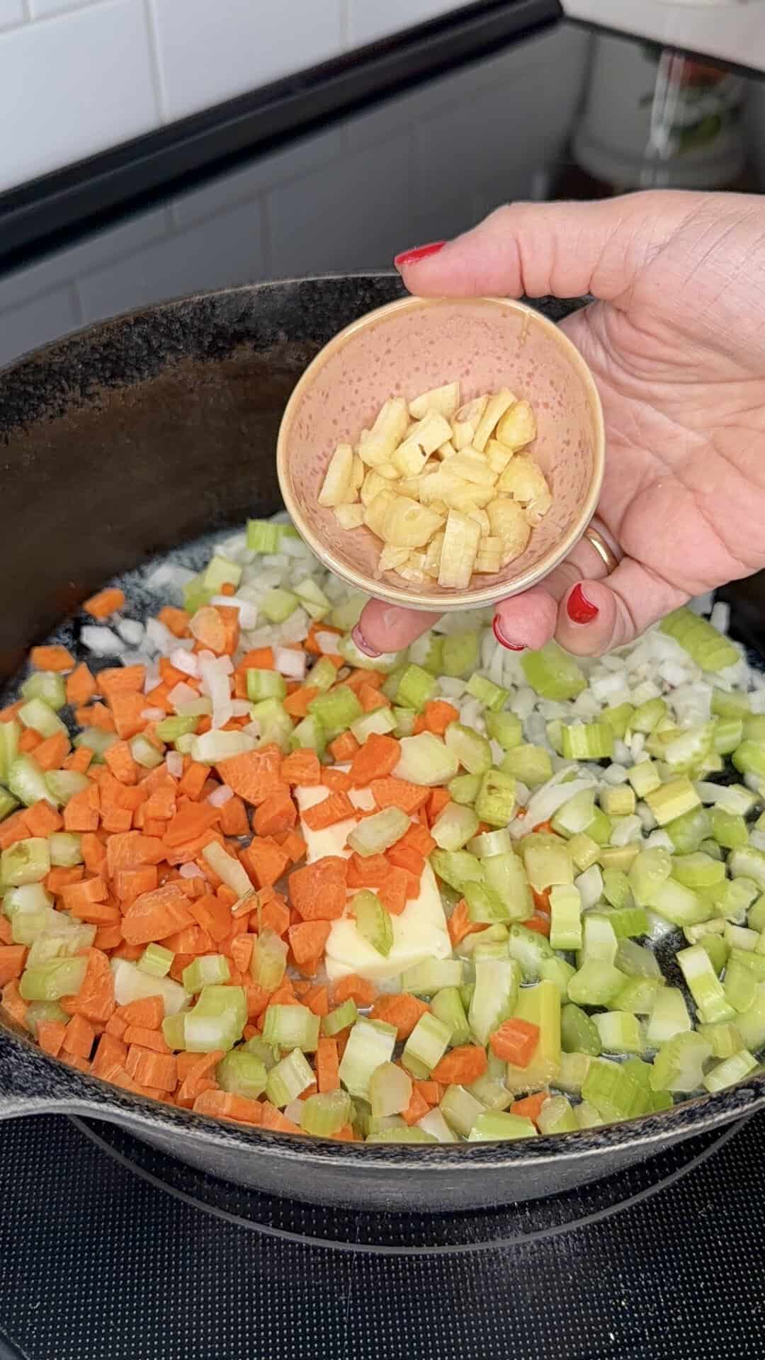 A hand holds a small bowl of chopped garlic over a pan with diced carrots, celery, and onions cooking on a stovetop.