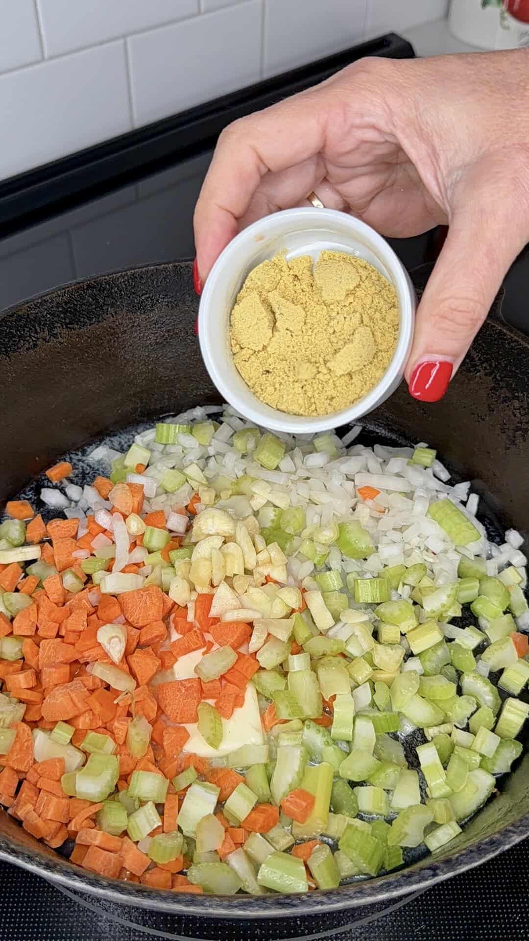 A hand holding a small bowl of nutritional yeast over a pan filled with chopped carrots, celery, onions, and garlic, ready to be cooked.