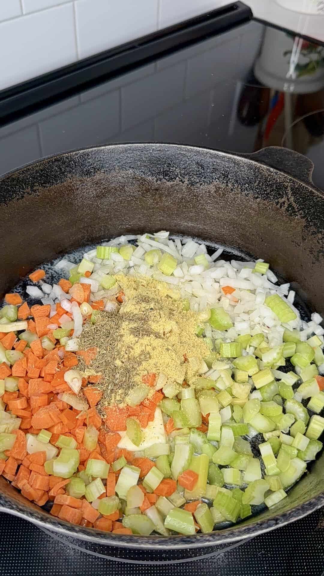 Chopped carrots, celery, and onions with seasonings sauté in a black cast iron skillet on a stovetop.