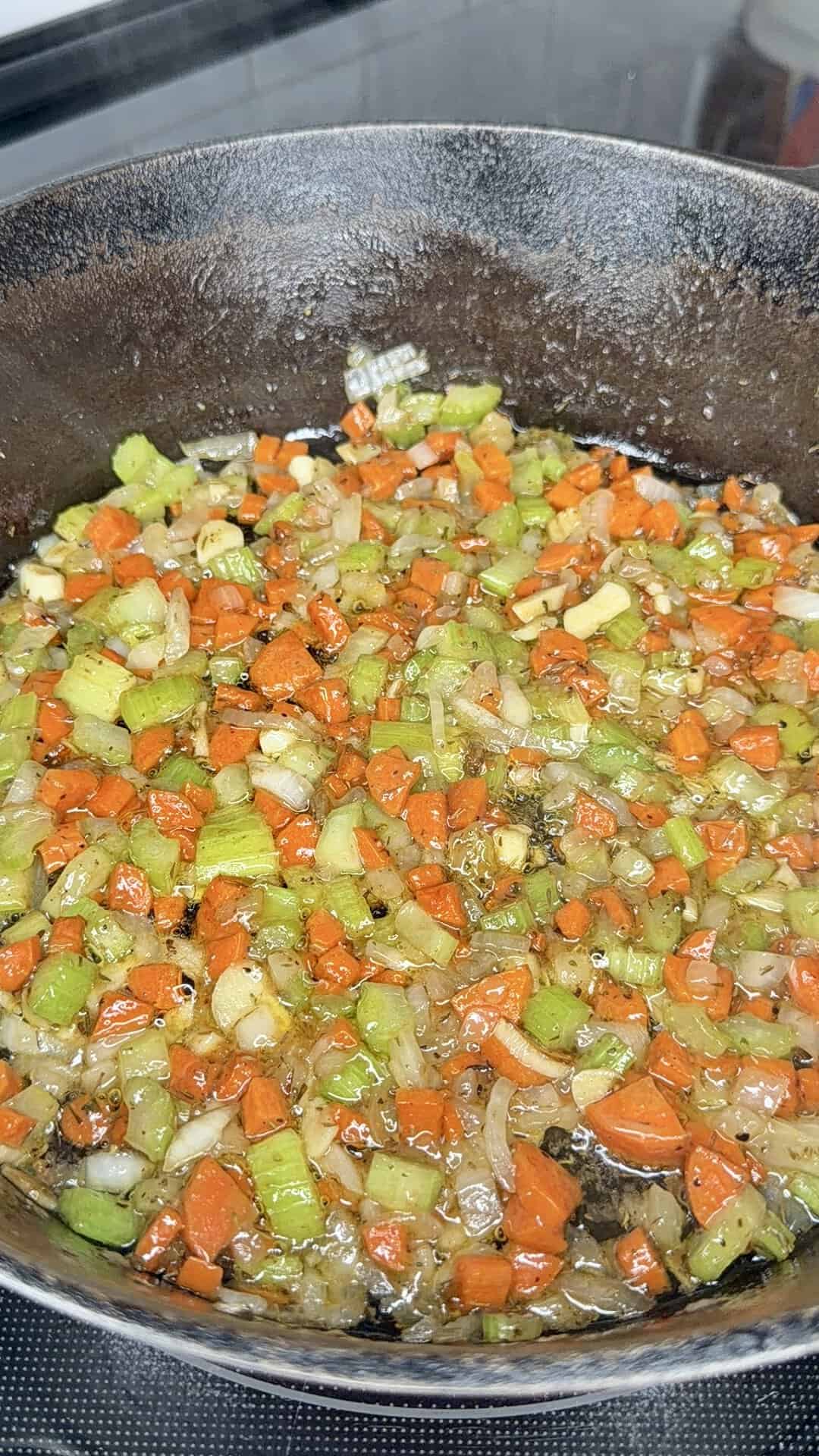 Diced carrots, celery, and onions sauté in oil in a black skillet on a stovetop, with visible herbs and seasonings mixed in.
