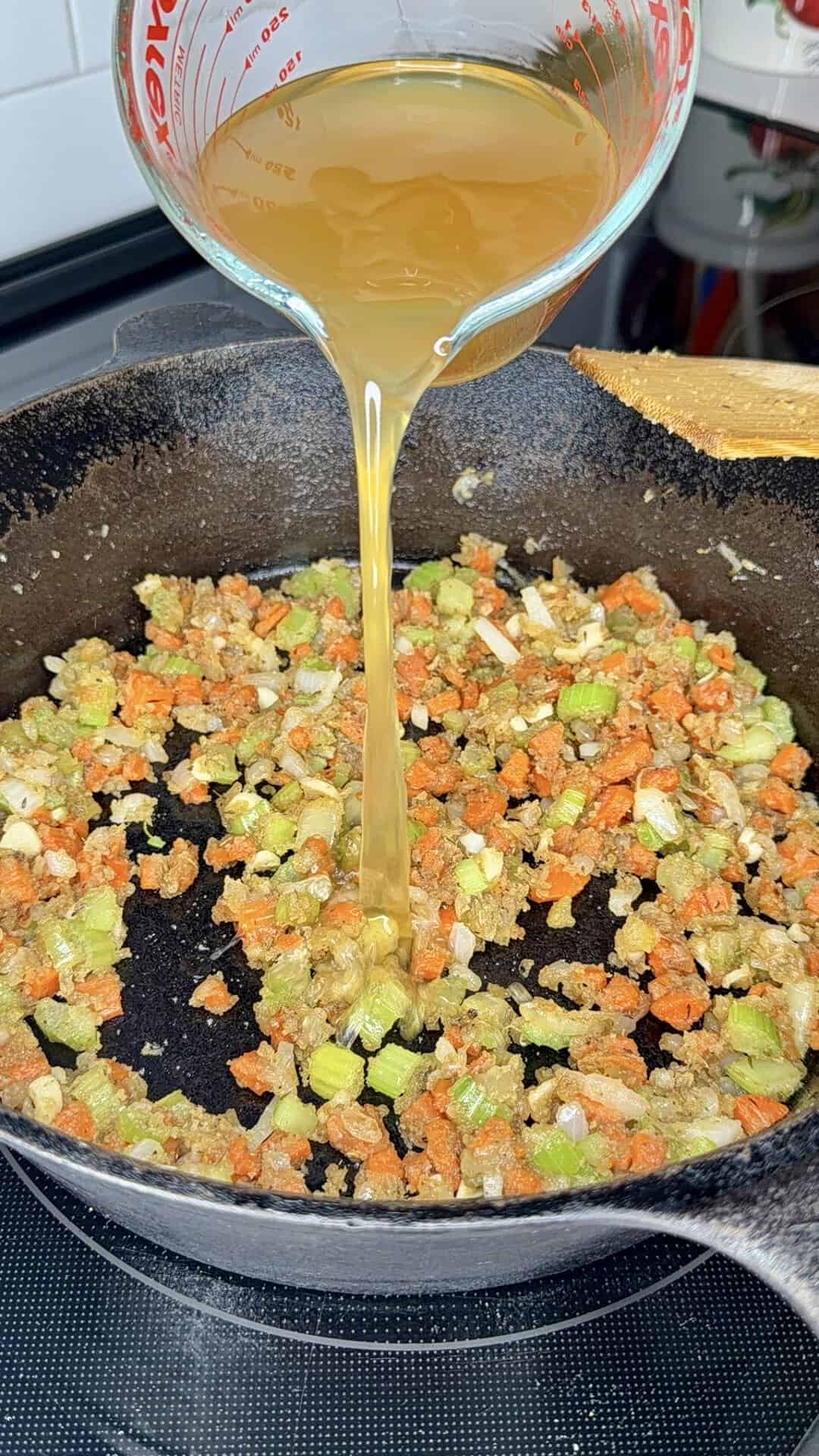 A measuring cup pours broth into a cast iron skillet with sautéed diced carrots, celery, and onions. A wooden spoon rests on the skillets edge.