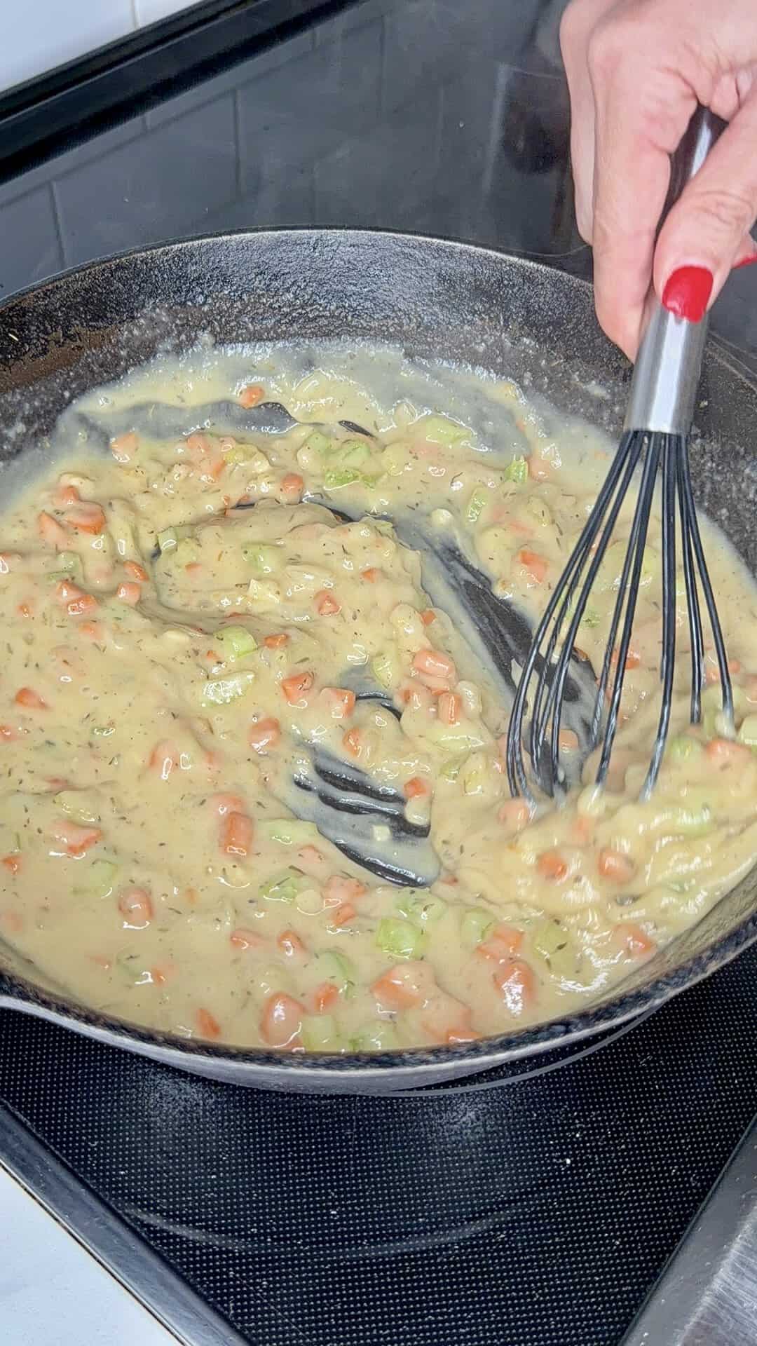 A hand whisks a creamy vegetable mixture with carrots and herbs in a black skillet on a stovetop.