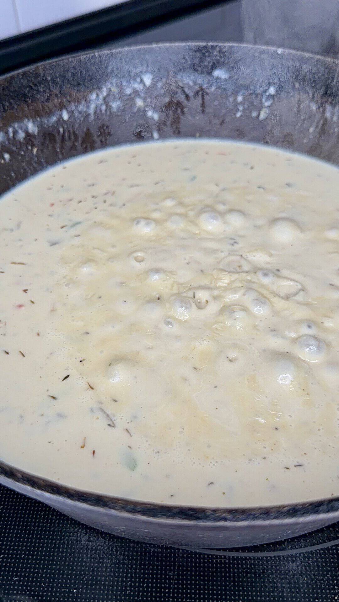 A close-up of a creamy soup or sauce boiling in a pot, with herbs and small ingredients visible in the mixture. Bubbles are forming on the surface as it heats on a stove.