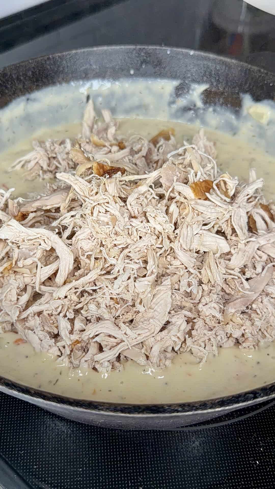 Shredded chicken sits in a skillet atop a creamy sauce, ready to be mixed together, on a stovetop.