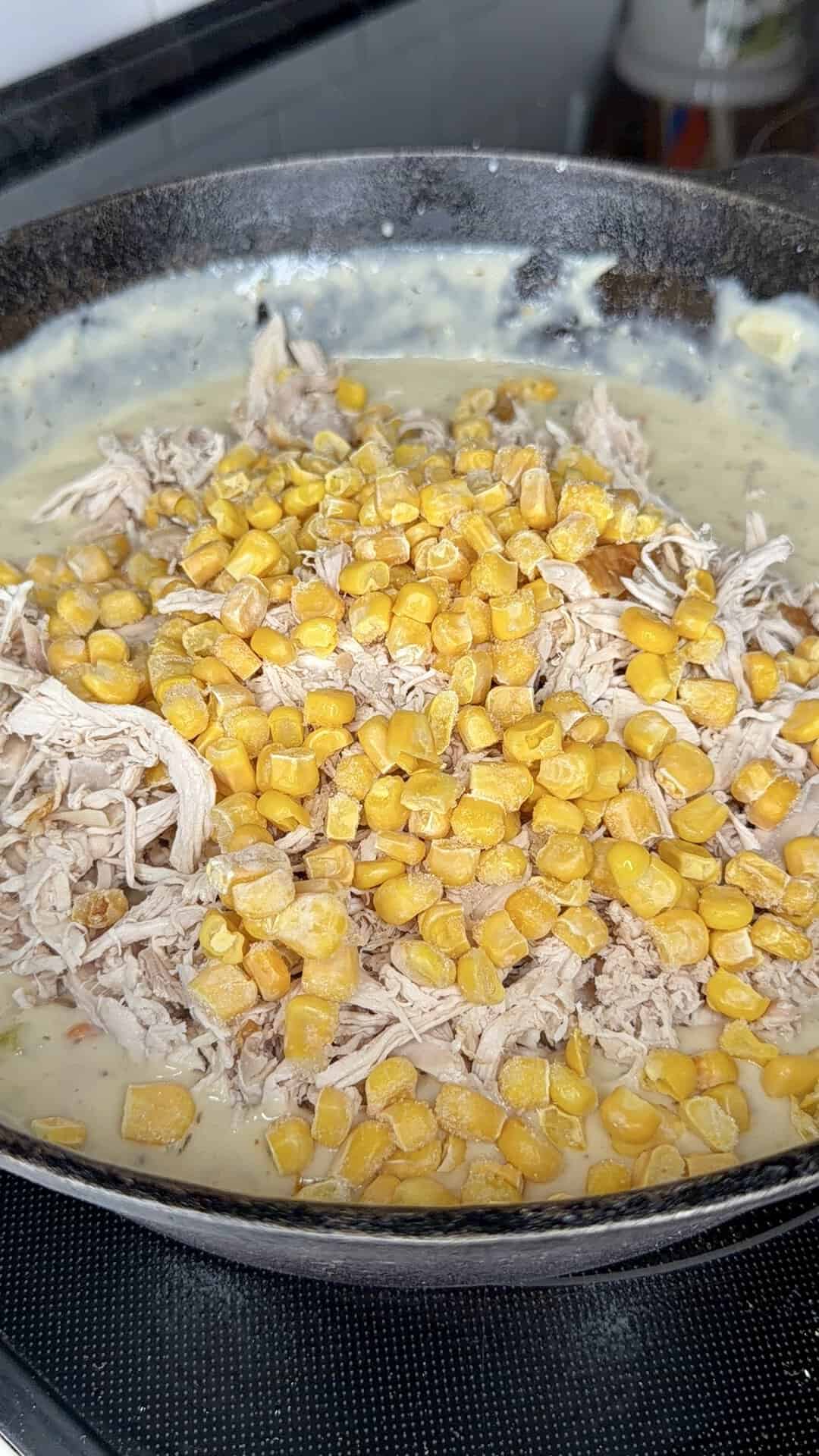 A close-up of a skillet filled with shredded chicken, yellow corn, and a creamy sauce cooking on a stovetop.