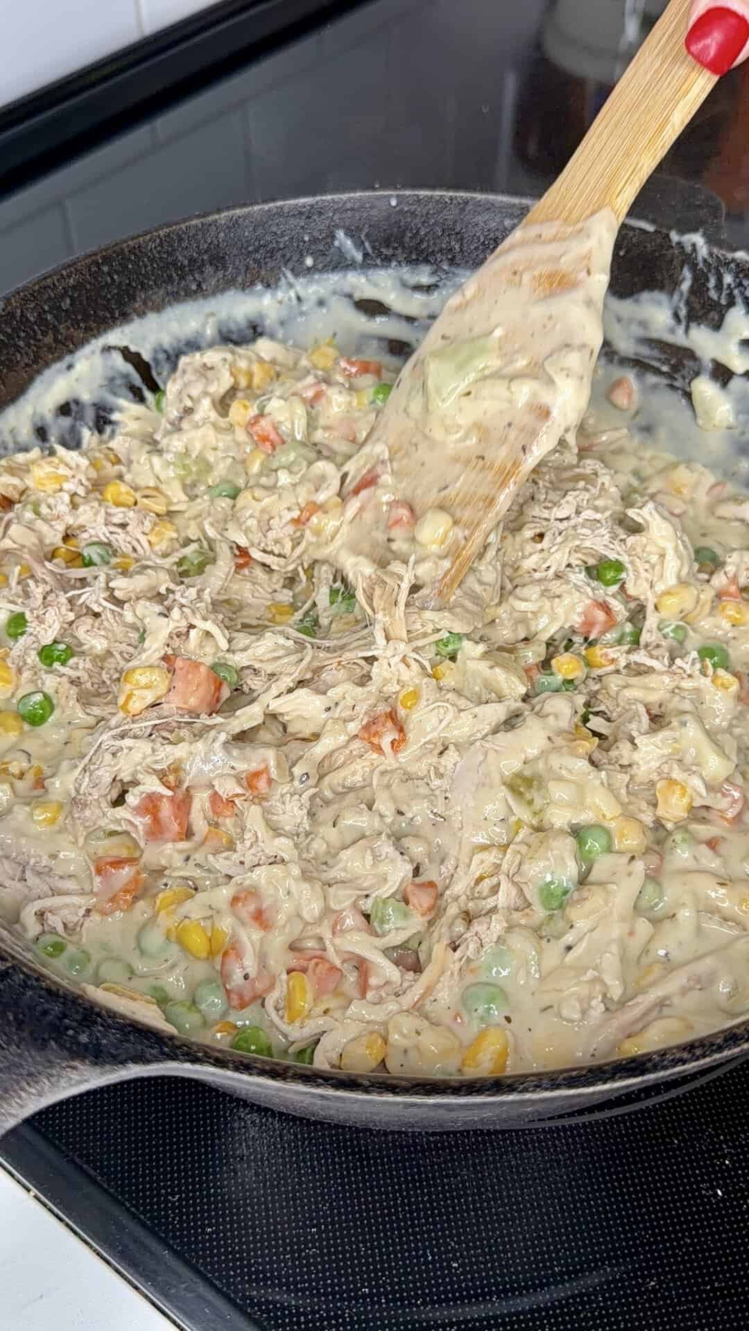 A hand stirs creamy shredded chicken with peas, corn, and carrots in a skillet on a stovetop.