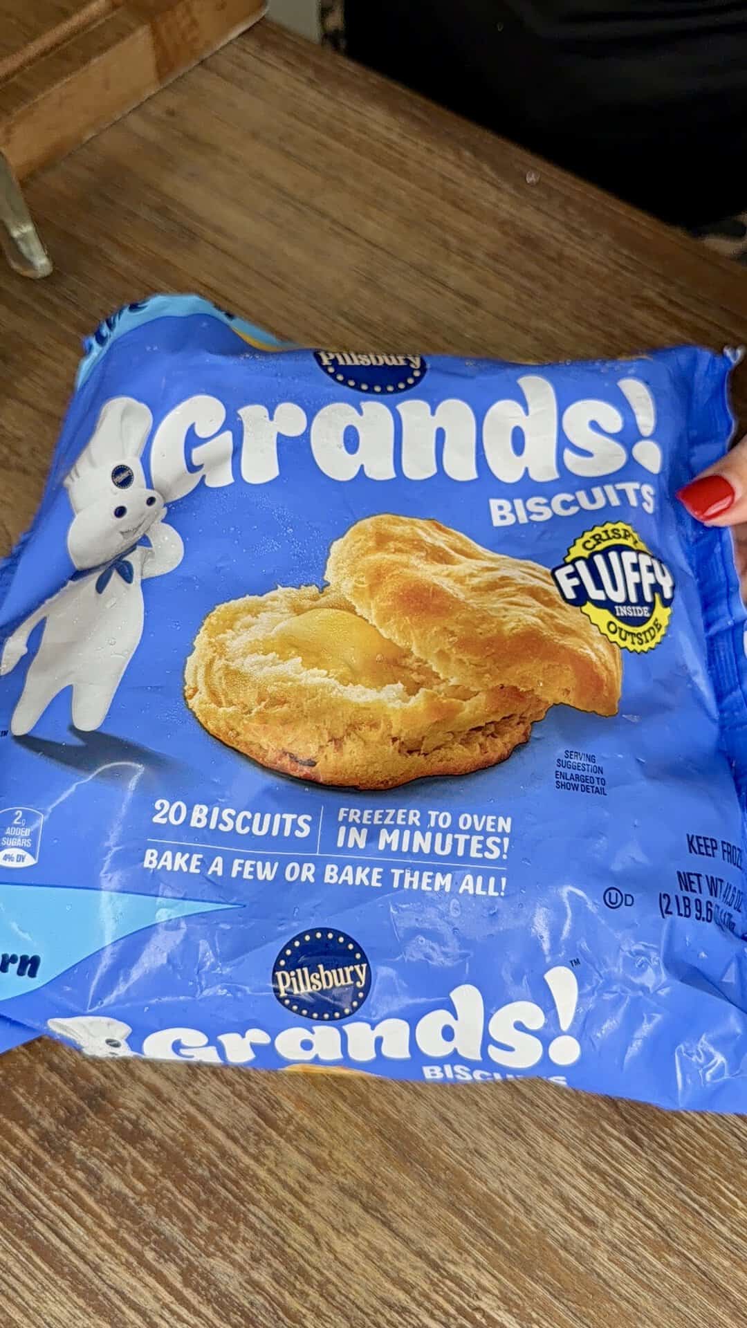 A hand holds a blue package of Pillsbury Grands! Biscuits, showing a biscuit and the Pillsbury Doughboy on the front. The package advertises 20 biscuits and instructions for baking.