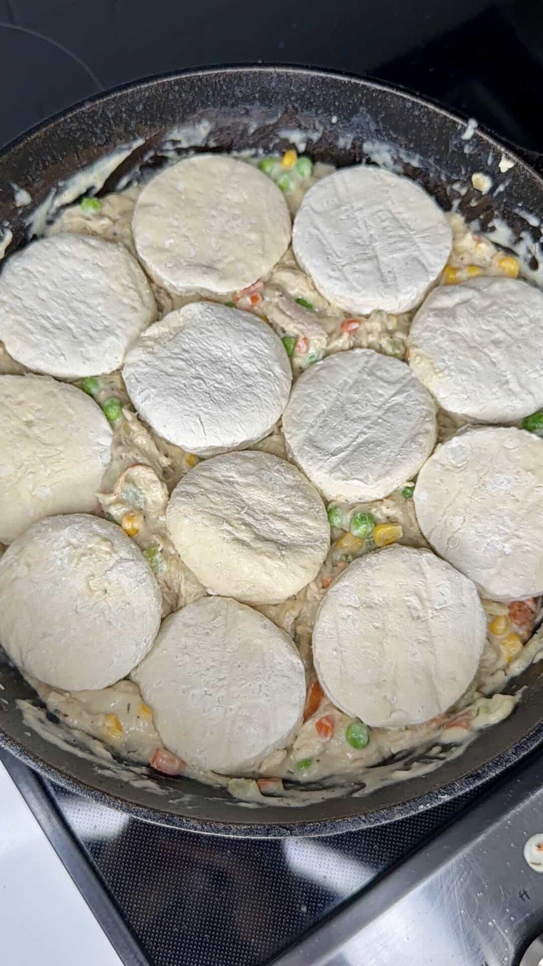 Unbaked biscuit dough rounds arranged on top of a creamy mixture with peas, corn, and carrots in a black skillet, ready to be baked.