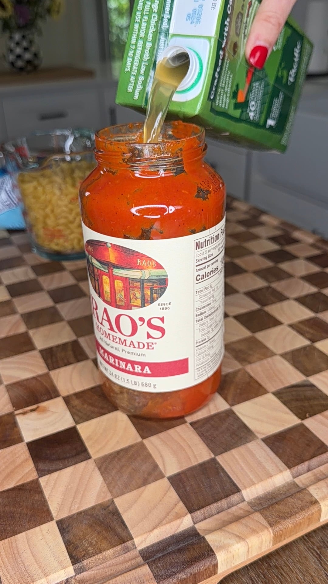 A hand pours liquid from a green carton into a jar of Raos Homemade Marinara sauce on a checkered wooden cutting board; dry pasta is in the background.