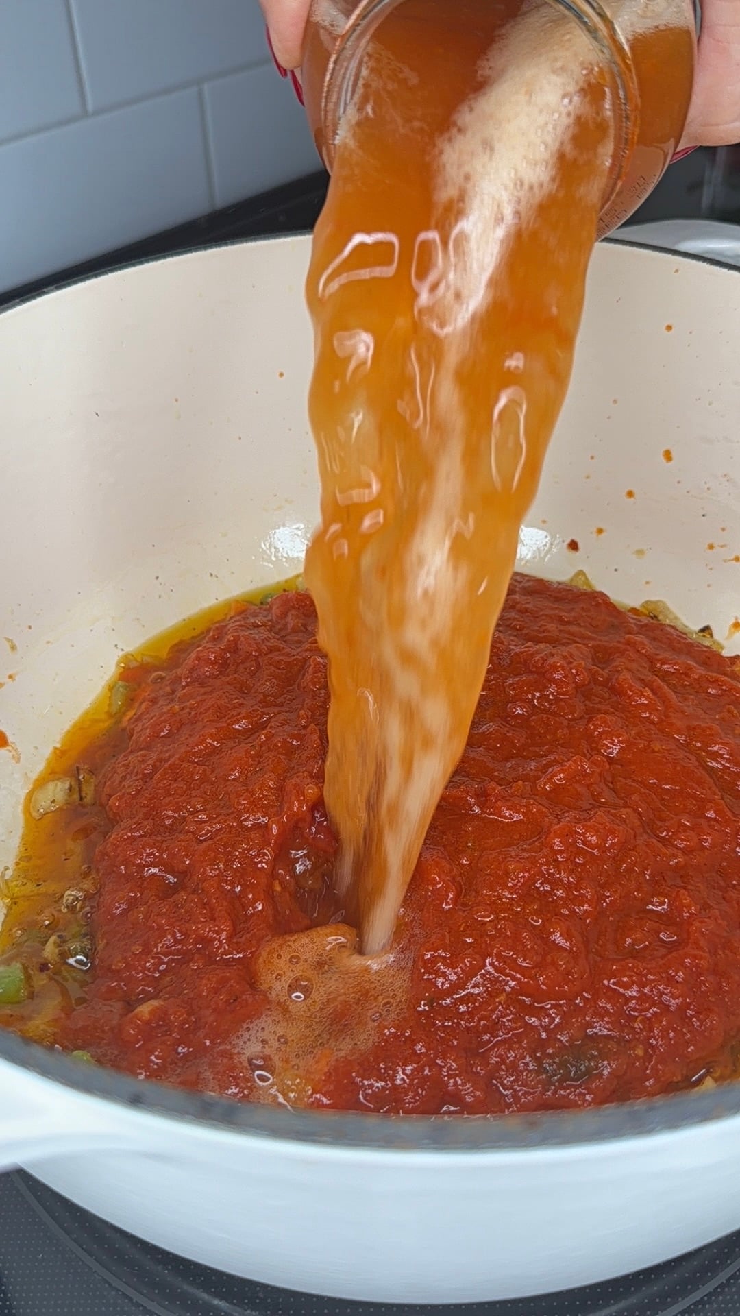 A hand pours a glass jar of liquid, likely water or broth, into a pot of red tomato sauce on a stovetop.
