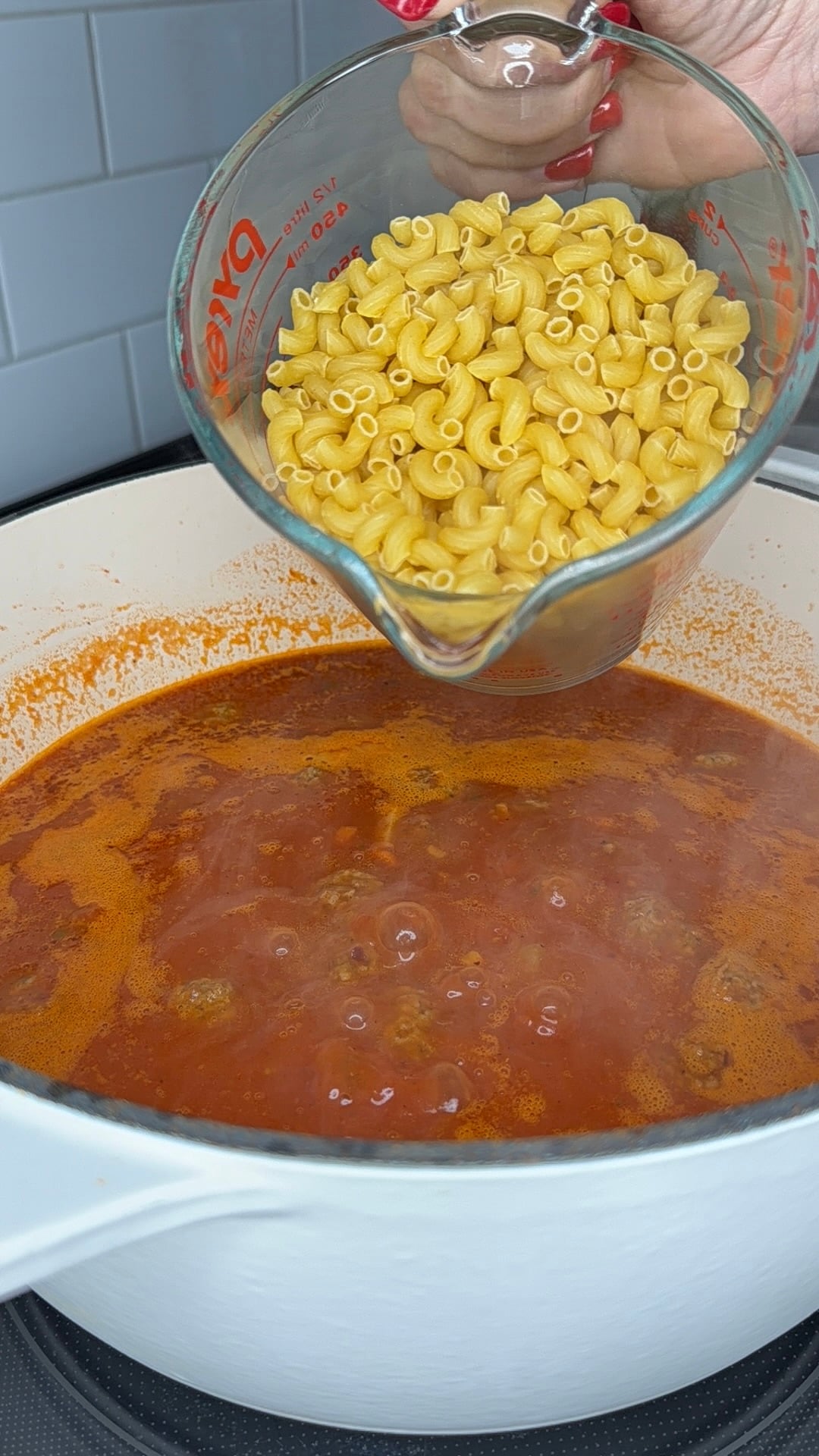 A hand holds a measuring cup of uncooked elbow macaroni above a pot of red soup or sauce on a stove, ready to pour the pasta in.