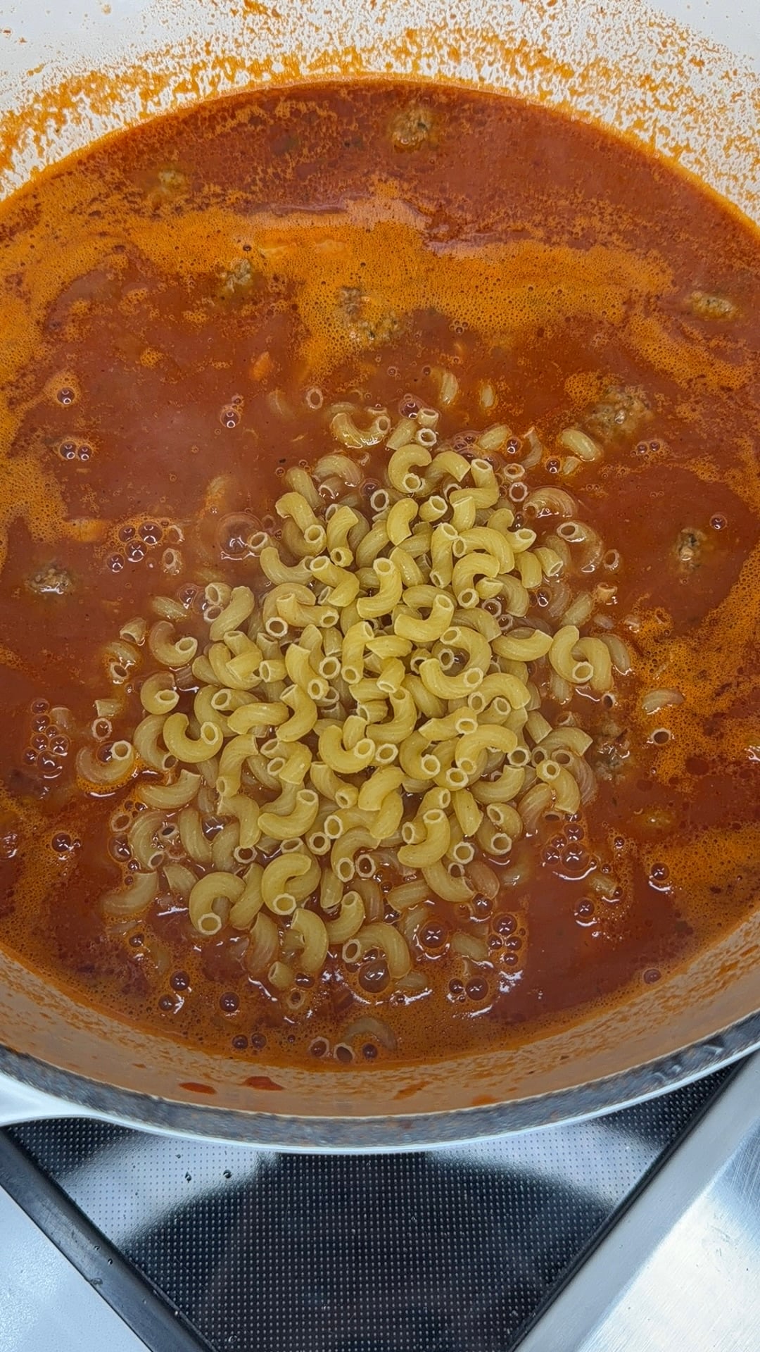 Uncooked elbow macaroni sits in the center of a pot filled with simmering red tomato sauce, ready to be stirred together on a stovetop.