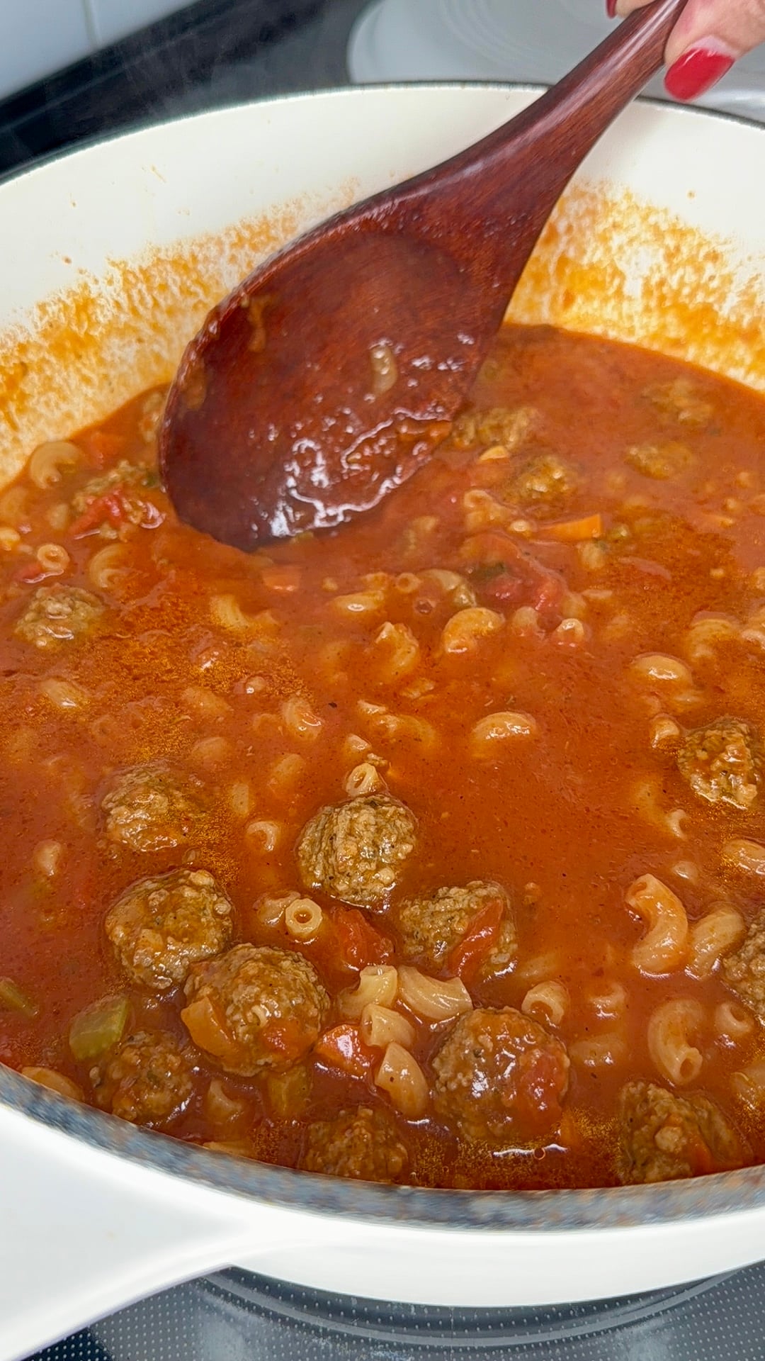 A hand stirs a pot of tomato-based soup with meatballs and elbow macaroni using a wooden spoon. The soup is simmering in a white pot on a stovetop.