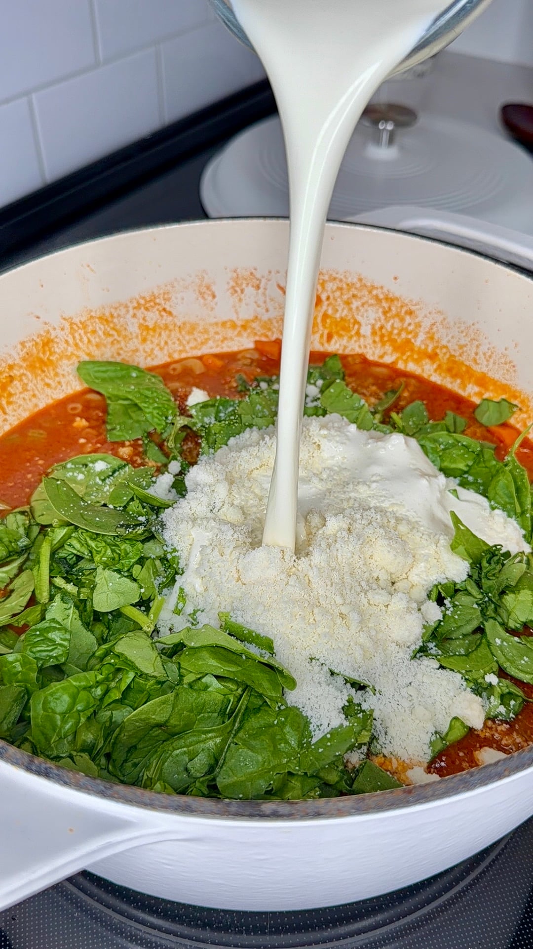 Milk being poured into a pot with tomato sauce, spinach, grated cheese, and cream, likely as ingredients for a creamy pasta dish.