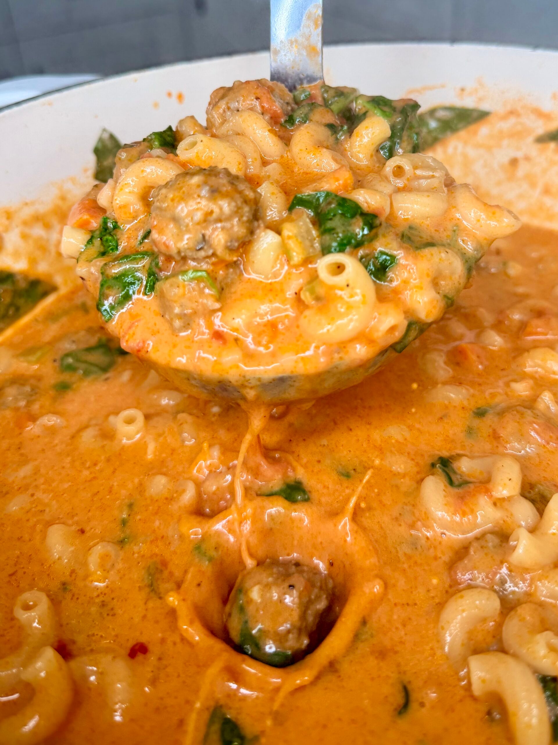 A ladle lifts creamy pasta with meatballs, spinach, and macaroni in a rich tomato sauce from a pot.