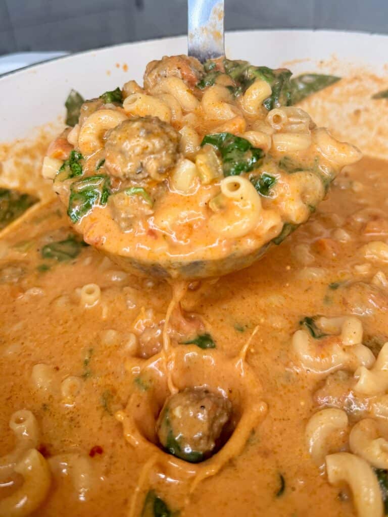 A ladle lifts creamy tomato-based pasta with meatballs, spinach, and elbow macaroni from a pot. The sauce is orange and cheesy, coating the pasta and ingredients.