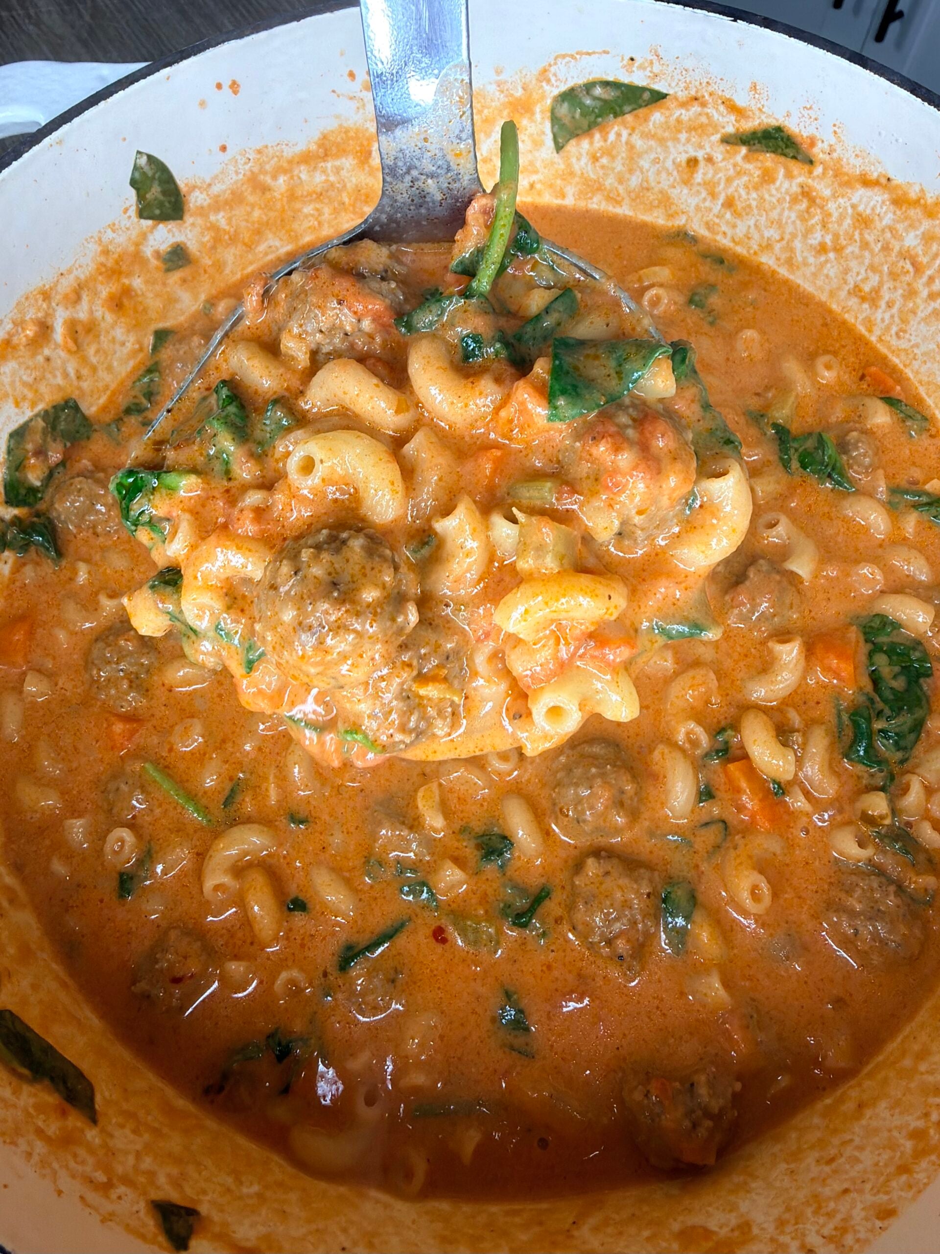 A ladle lifts creamy tomato pasta soup with meatballs, macaroni, spinach, and chunks of tomato from a large pot.