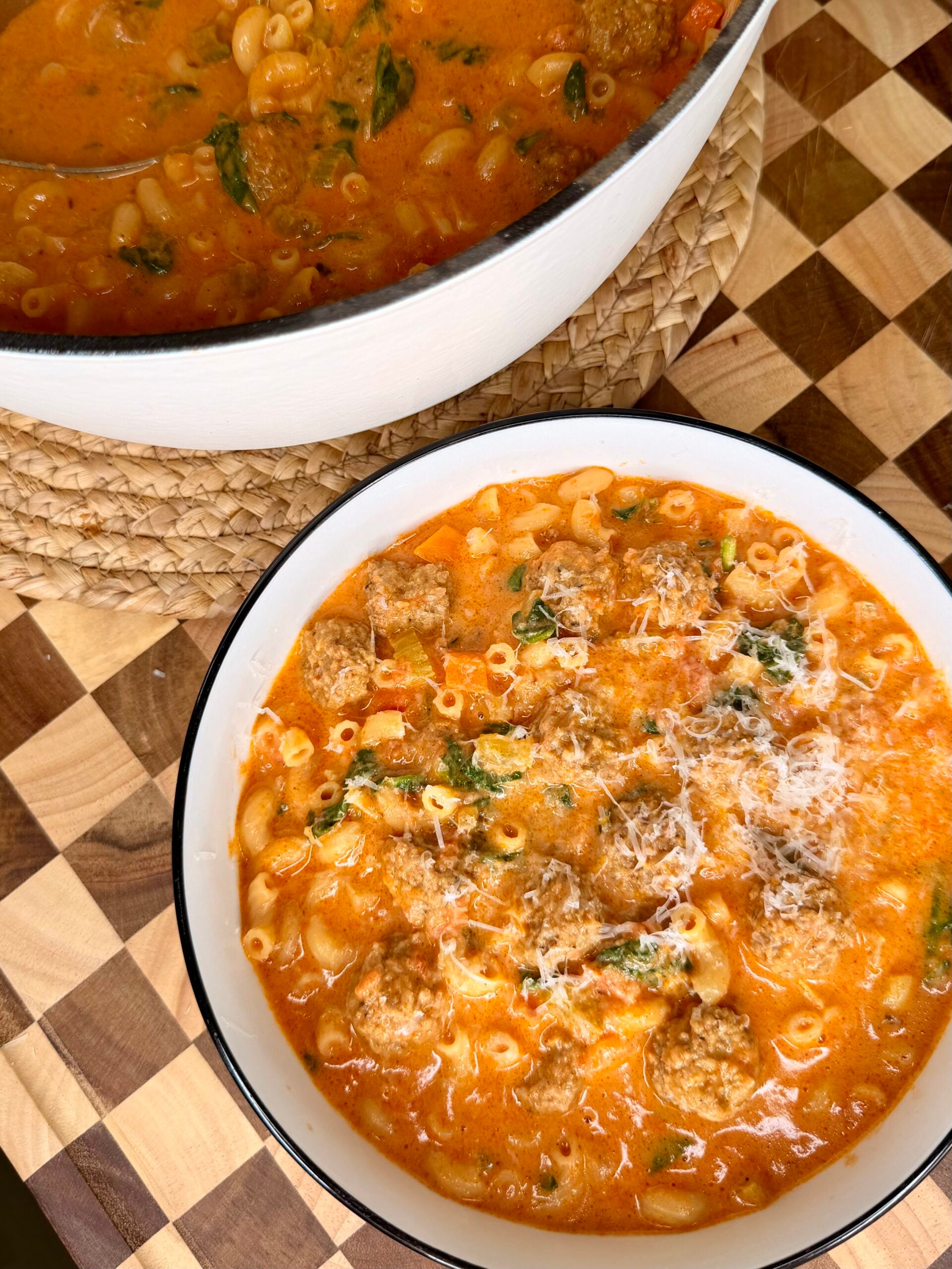 A bowl of creamy tomato soup with macaroni, meatballs, spinach, and grated cheese sits next to a large pot of the same soup on a wooden checkered table.