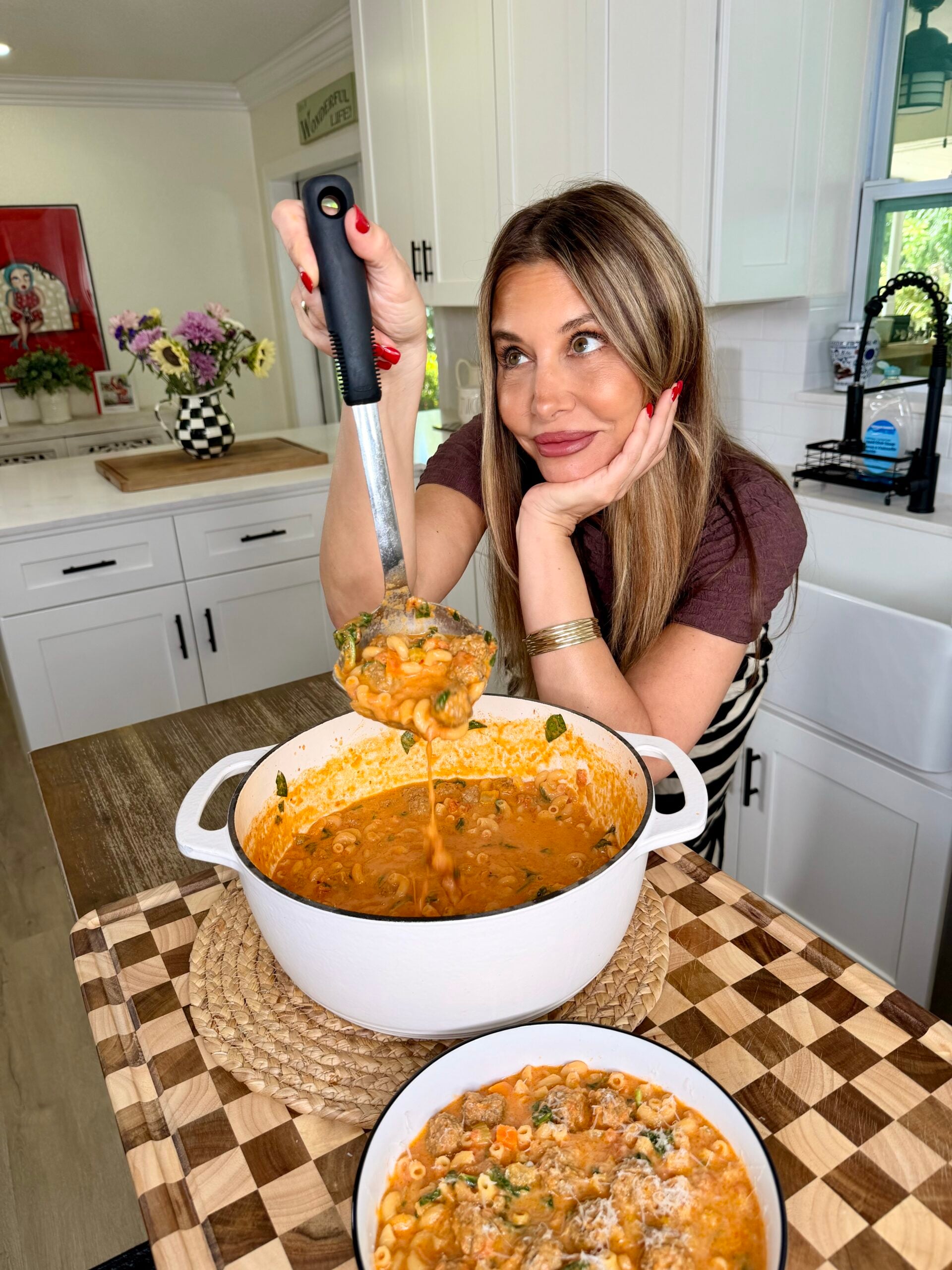 A woman in a kitchen serves pasta soup from a white pot into a bowl, smiling and resting her chin on her hand. The kitchen is bright with white cabinets and flowers in the background.