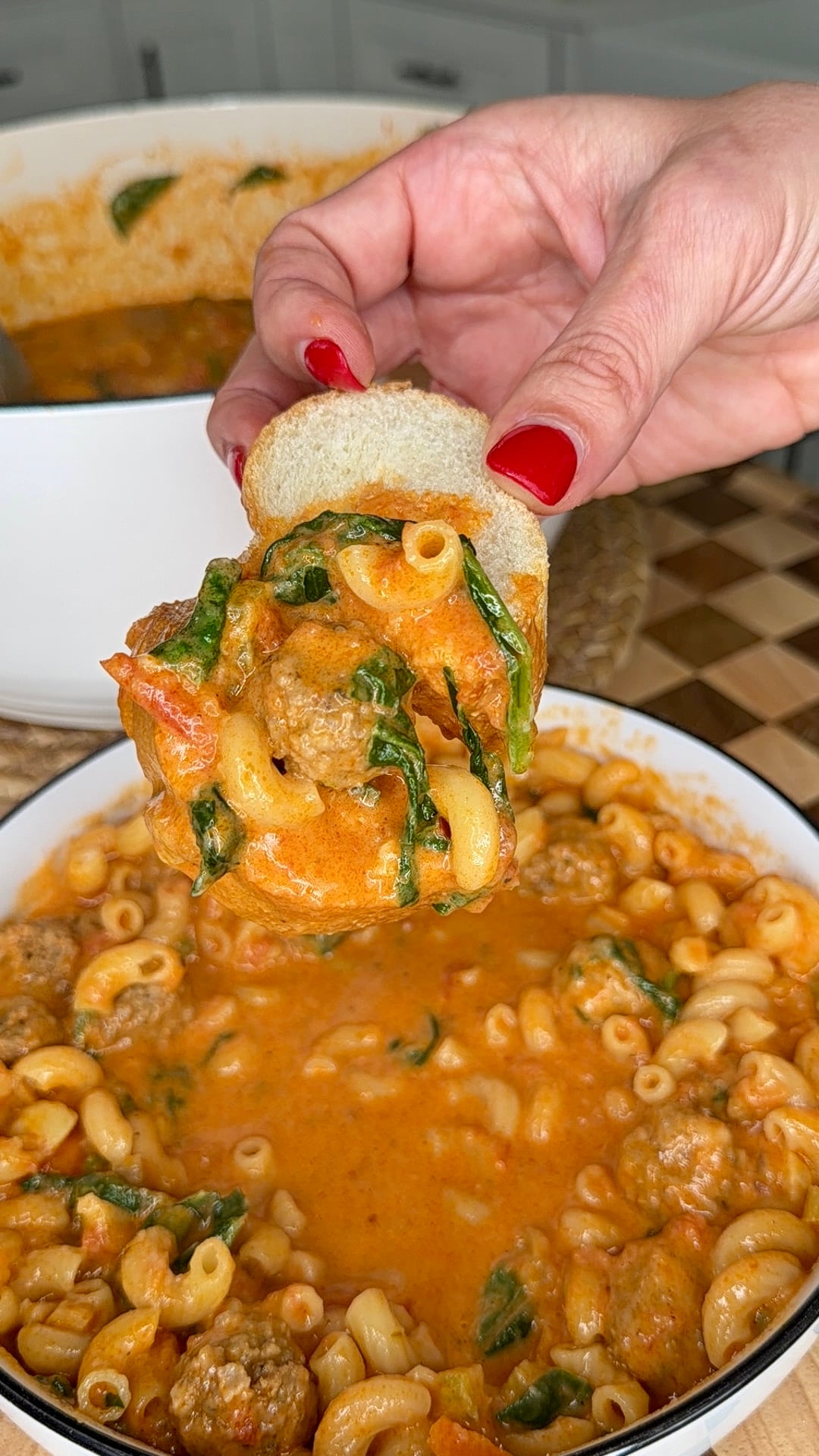 A hand with red nails holds bread scooping cheesy pasta, meatballs, spinach, and tomato sauce from a white bowl filled with macaroni pasta. A pot of more pasta sits in the background.