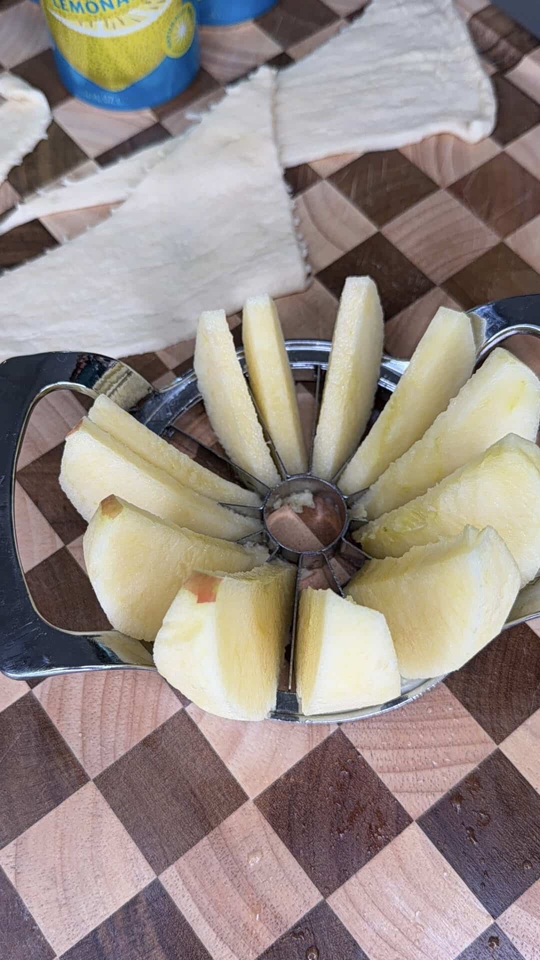 An apple slicer tool holding a freshly sliced apple sits on a checkerboard patterned cutting board. In the background, there is a blue can and pieces of raw pastry dough, suggesting preparation for baking.