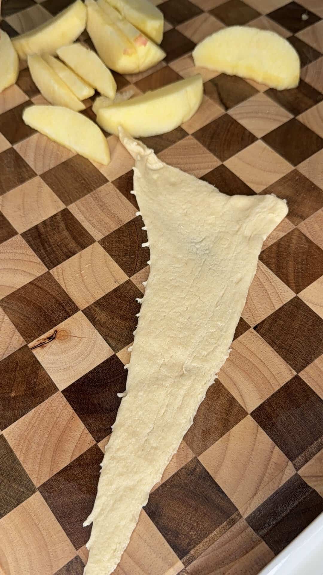 A triangle of uncooked dough and sliced apple pieces are placed on a checkered wooden cutting board.