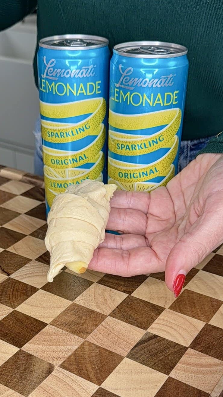 A hand with red nail polish holds an uncooked croissant dough in front of two blue cans of Lemonati Sparkling Lemonade on a checkered wooden surface.