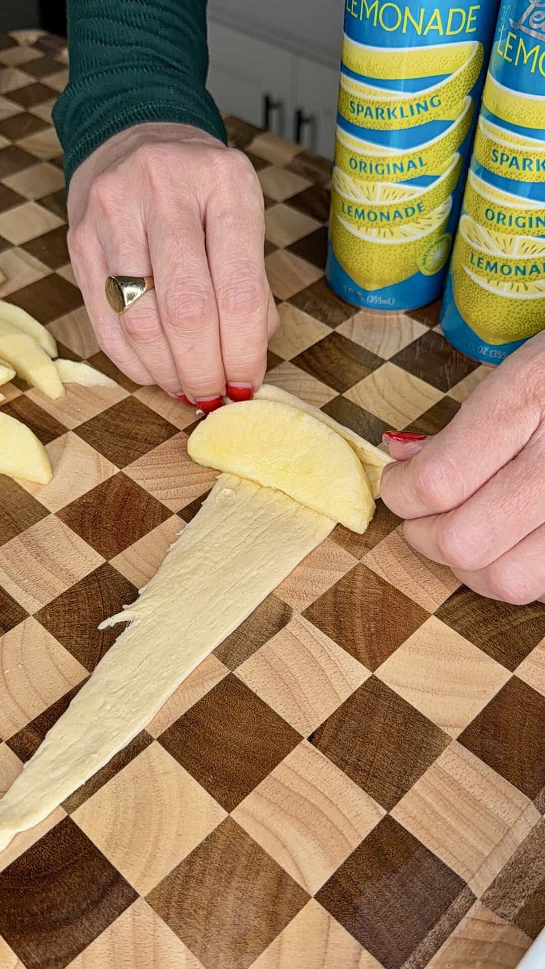 A person with manicured nails folds a slice of apple into a triangular piece of pastry dough on a wooden checkerboard cutting board, with two cans of sparkling lemonade in the background.