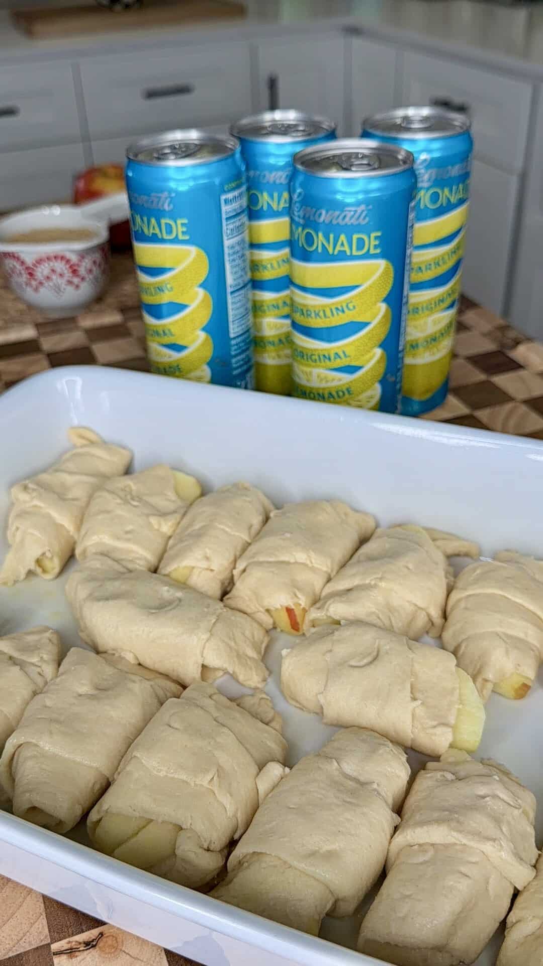 A white dish with unbaked crescent rolls, some showing apple slices inside, sits on a checkered counter. Behind it are four cans of lemon sparkling lemonade. Kitchen items are in the background.