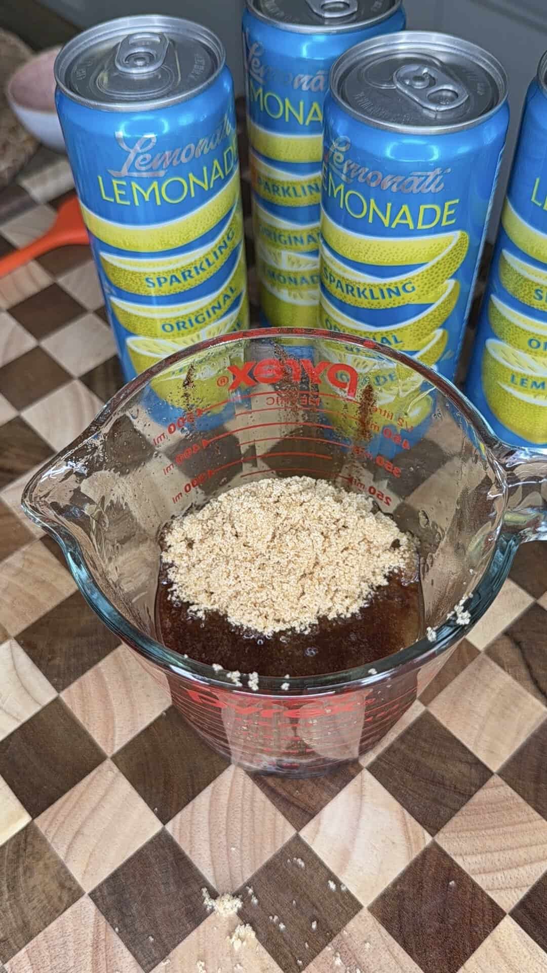 A glass measuring cup with brown liquid and brown sugar on top sits on a checkered cutting board. Behind it are four tall cans of sparkling lemonade.