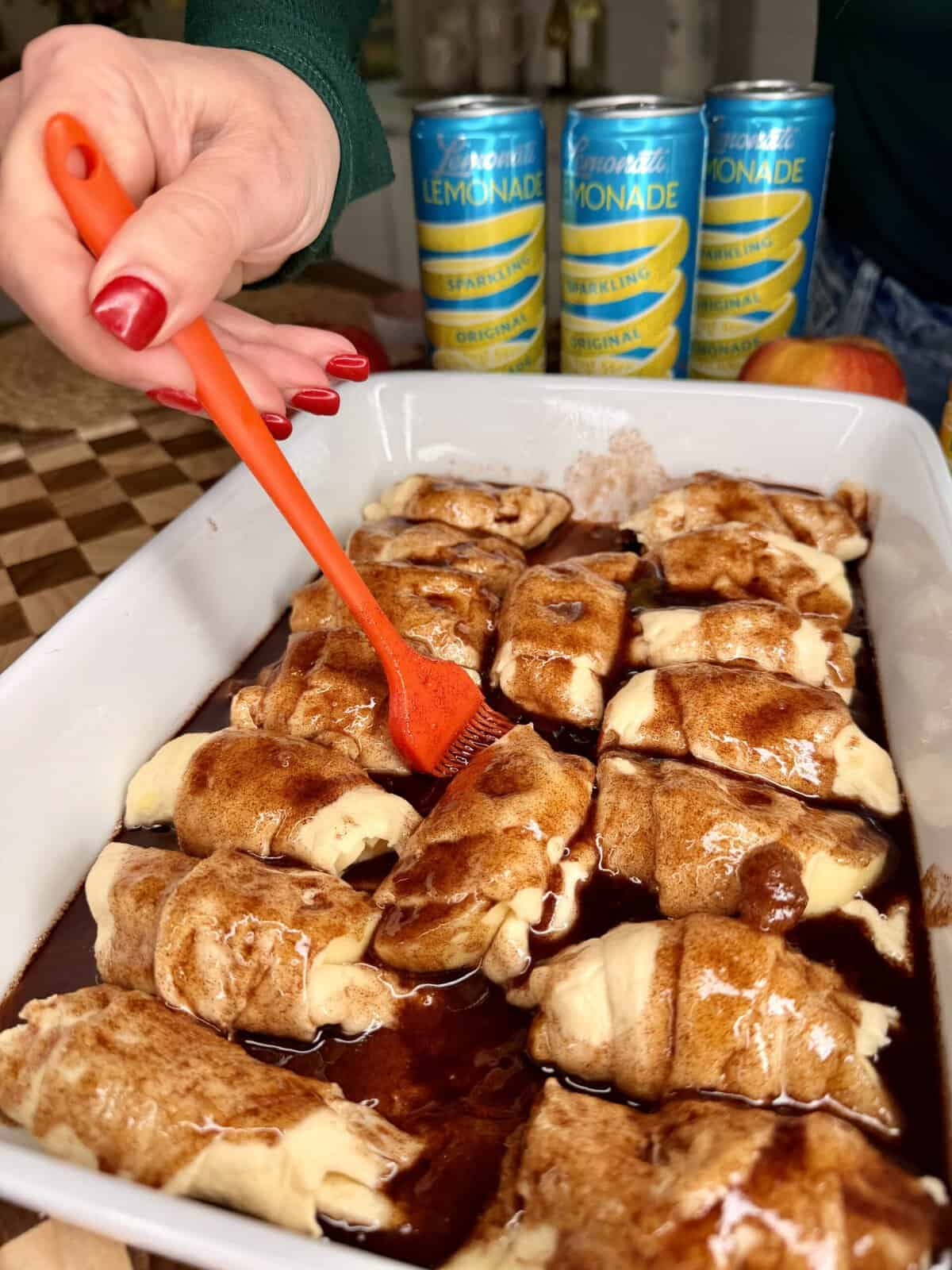 A hand uses an orange brush to spread sauce over crescent rolls in a baking dish. Three cans of lemonade are in the background.