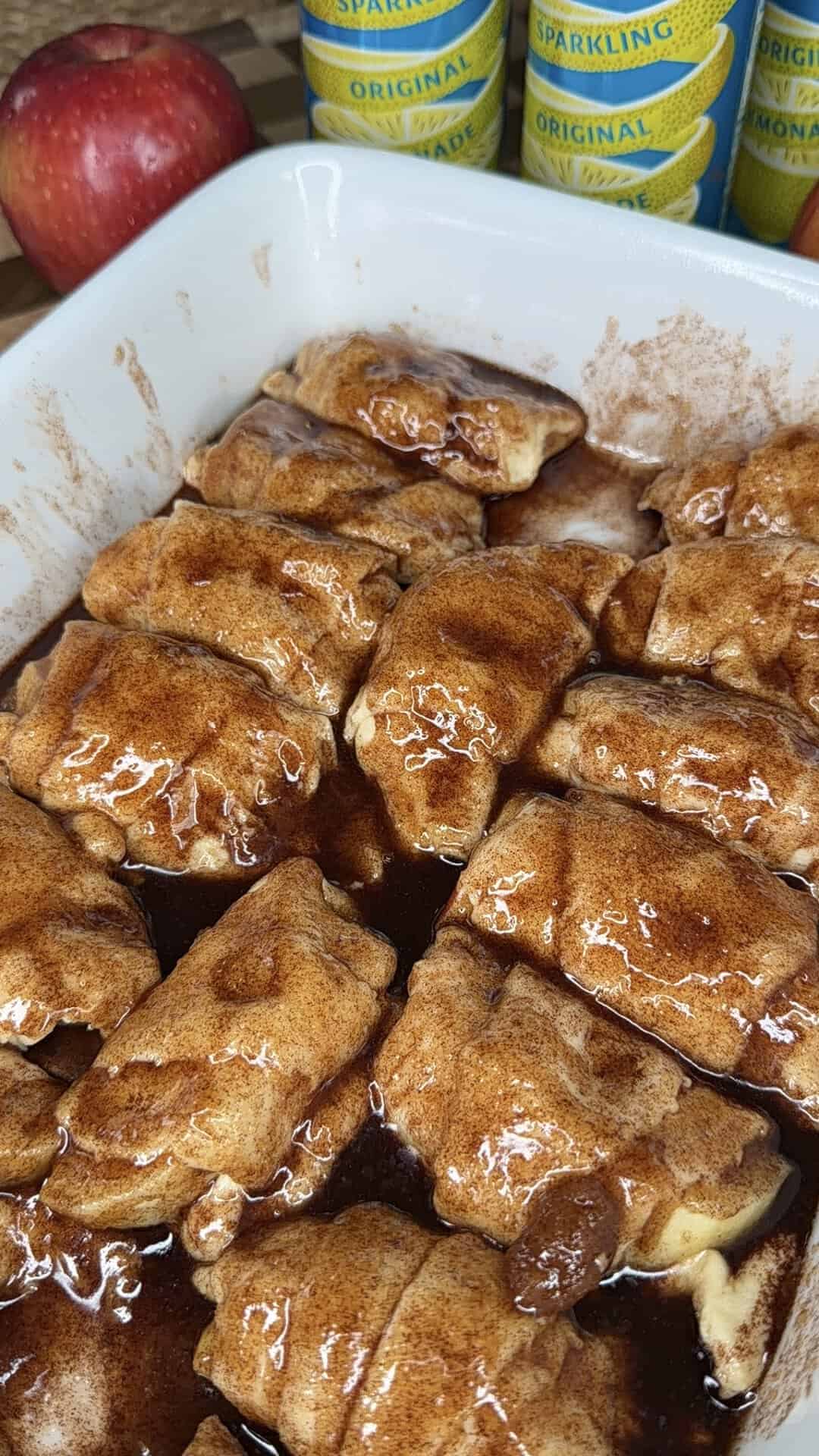 Golden-brown baked pastries in a white dish are drizzled with cinnamon glaze, with sparkling water cans and a red apple in the background.