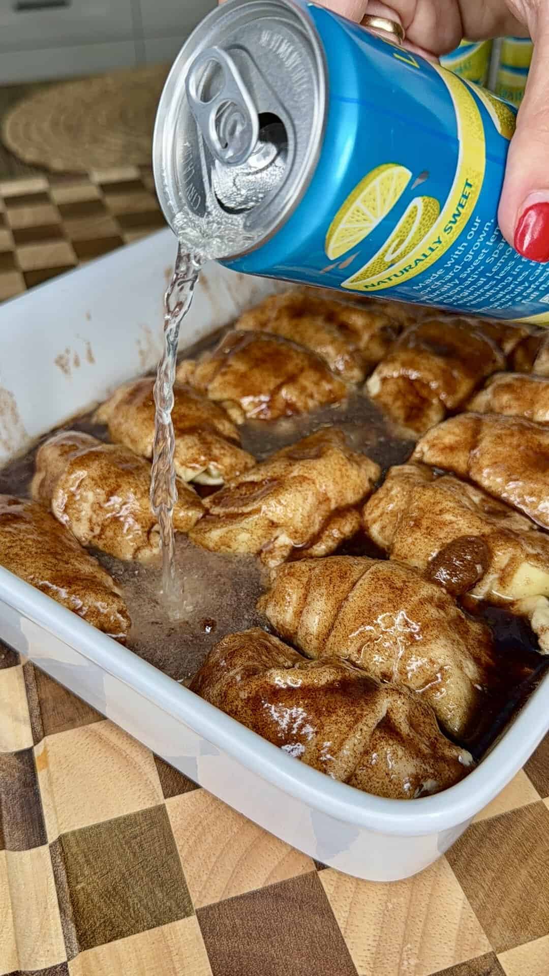 A hand pours clear soda from a blue can over raw cinnamon rolls in a white baking dish on a checkered wooden surface.