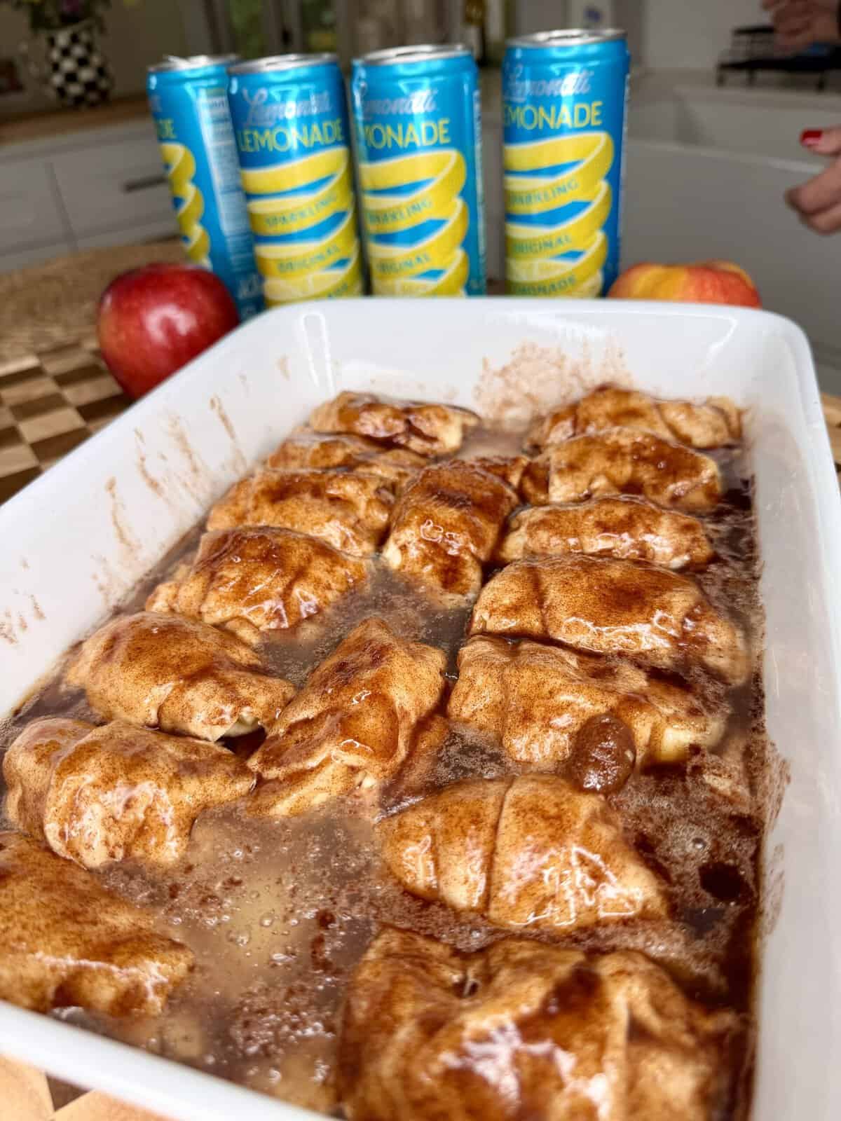 A white dish filled with baked, golden-brown cinnamon rolls, with bubbling sauce. In the background, there are four cans of lemonade, two apples, and part of a hand on a checkered countertop.