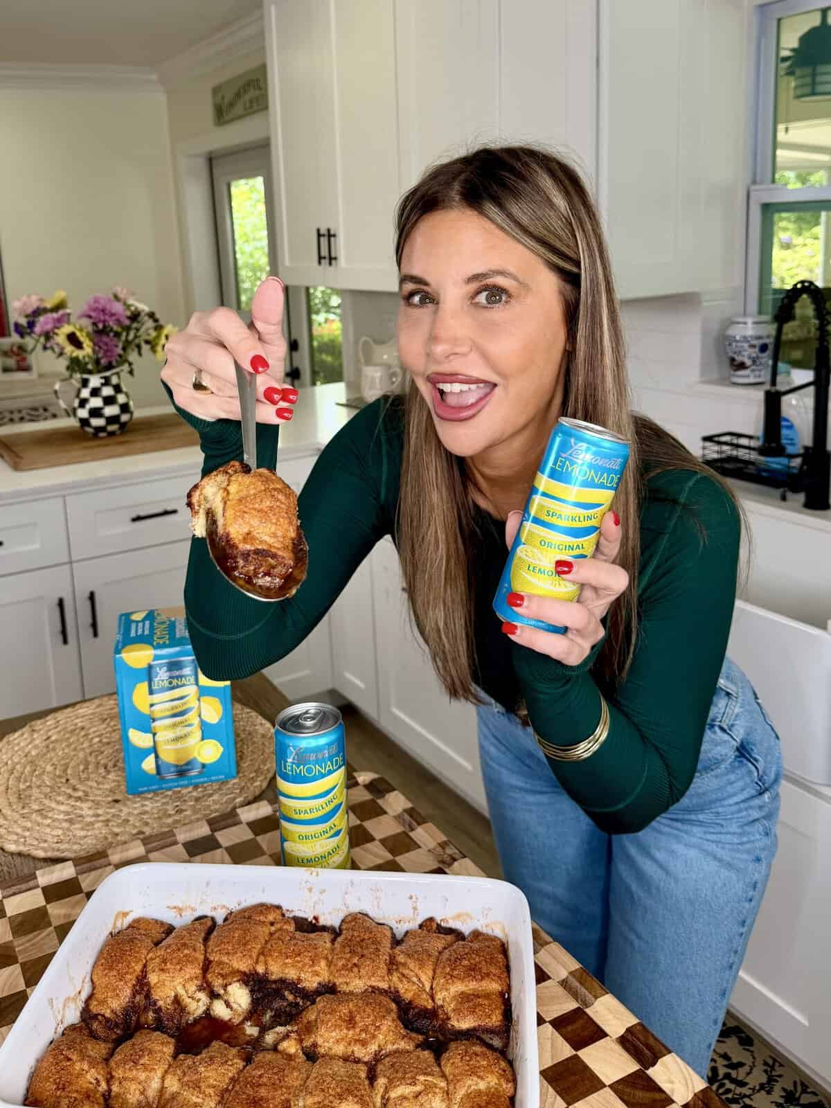 A smiling woman in a green top holds a can of sparkling lemonade in one hand and lifts a piece of baked dessert from a white dish in a bright kitchen.