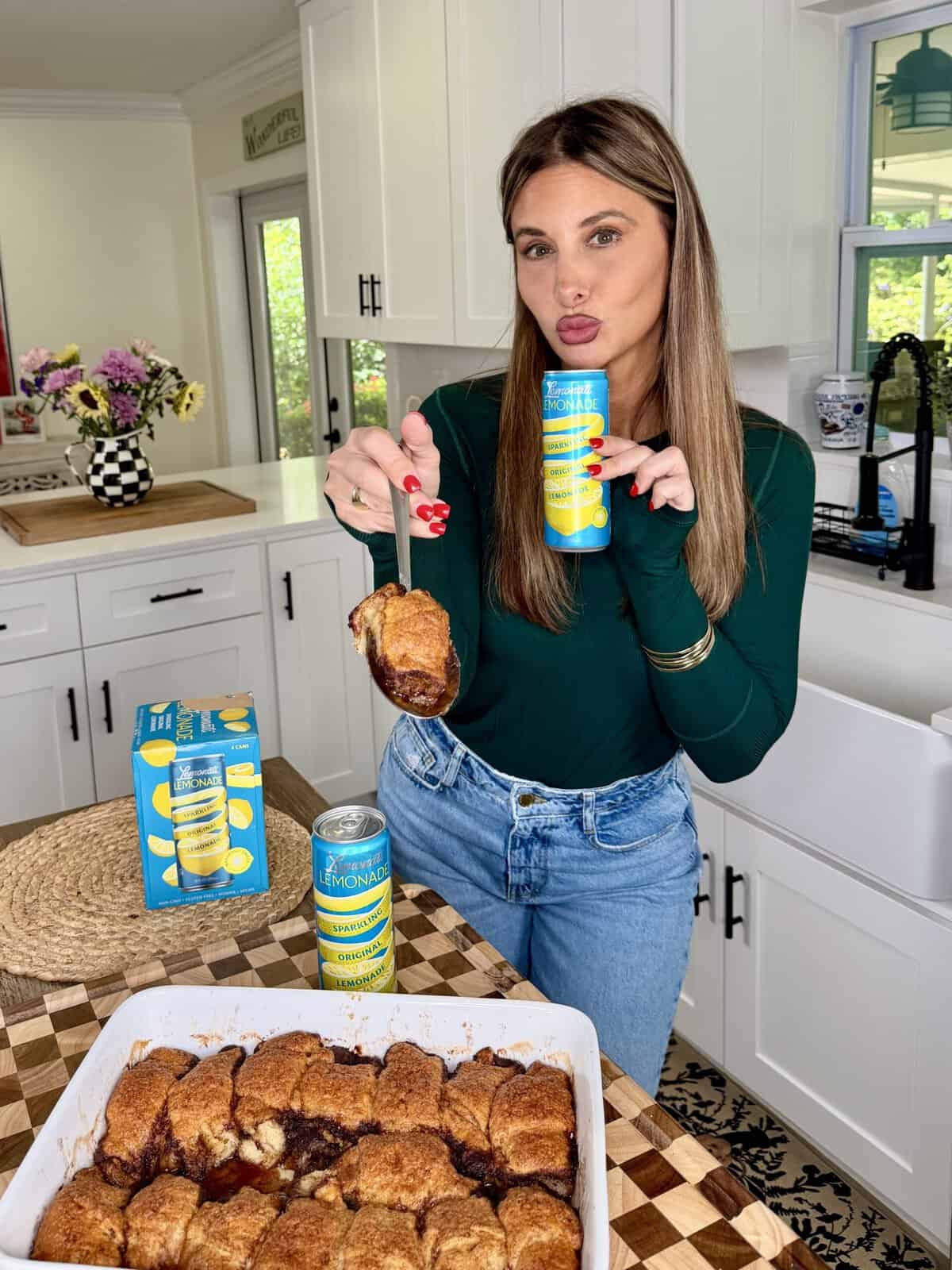 A woman in a green top and jeans holds a can of lemonade and a serving of baked dessert in a modern kitchen, with a baking dish and lemonade cans on the counter.