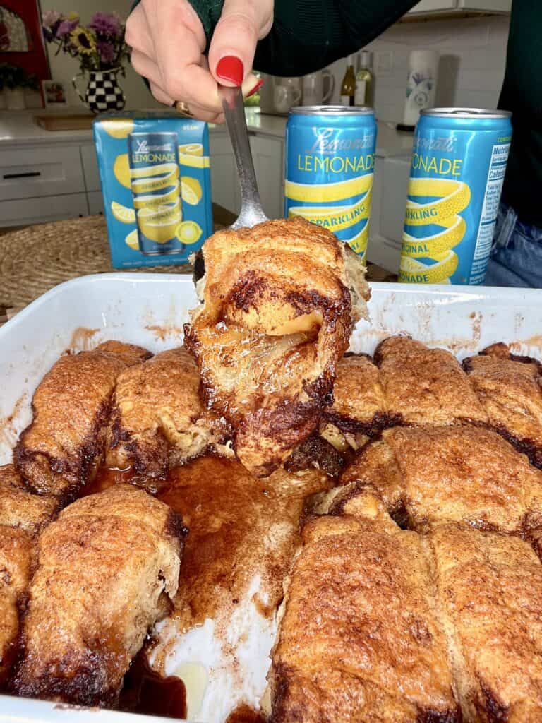A hand with red nail polish lifts a serving of cinnamon sugar baked rolls from a white dish. Three cans of sparkling lemonade and a box are visible in the background on a kitchen counter.