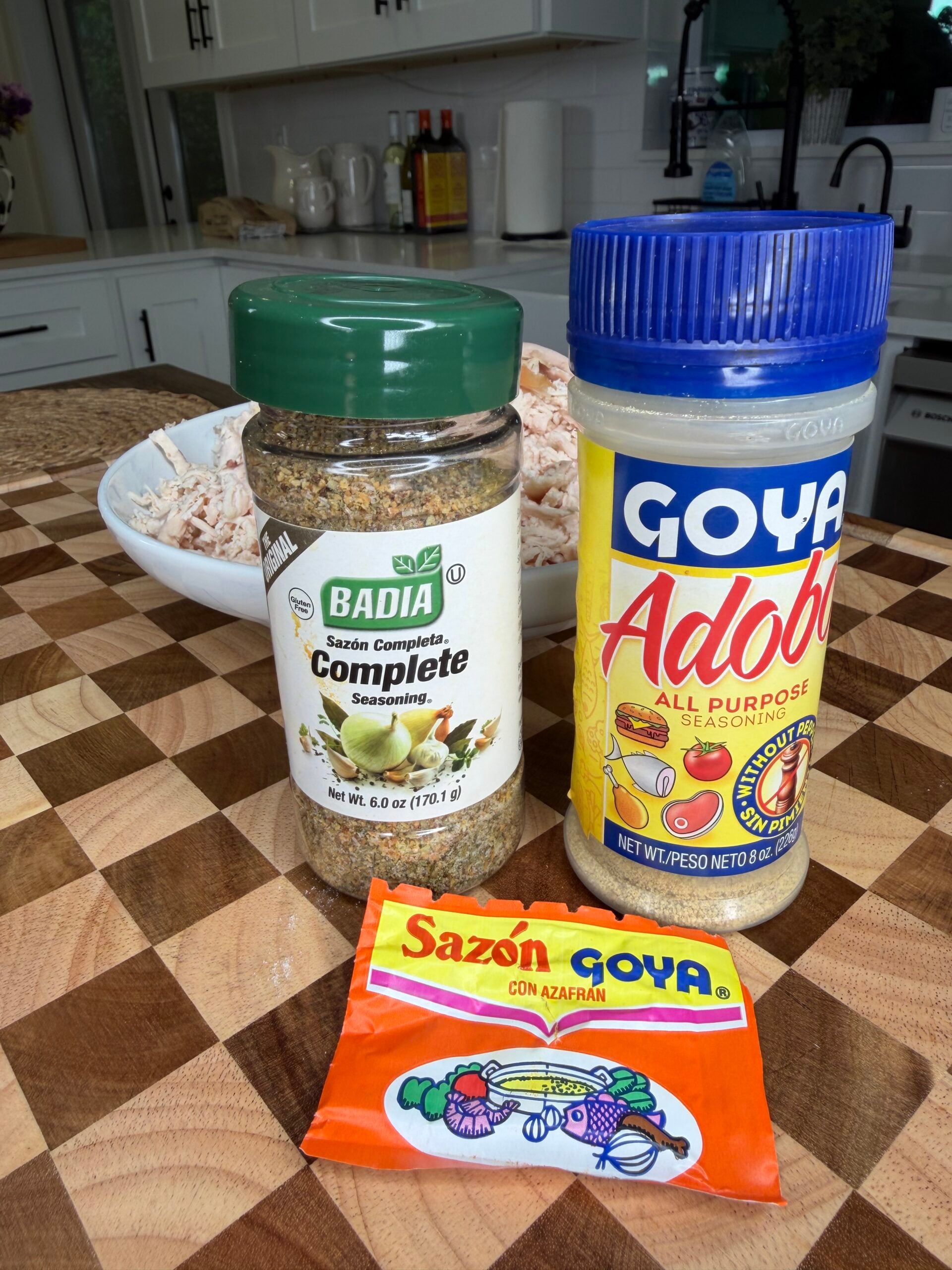 Three seasoningsโBadia Sazon Complete, Goya Adobo, and a packet of Sazon Goya con Azafrรกnโon a wooden cutting board, with a bowl of shredded chicken in the background in a kitchen setting.
